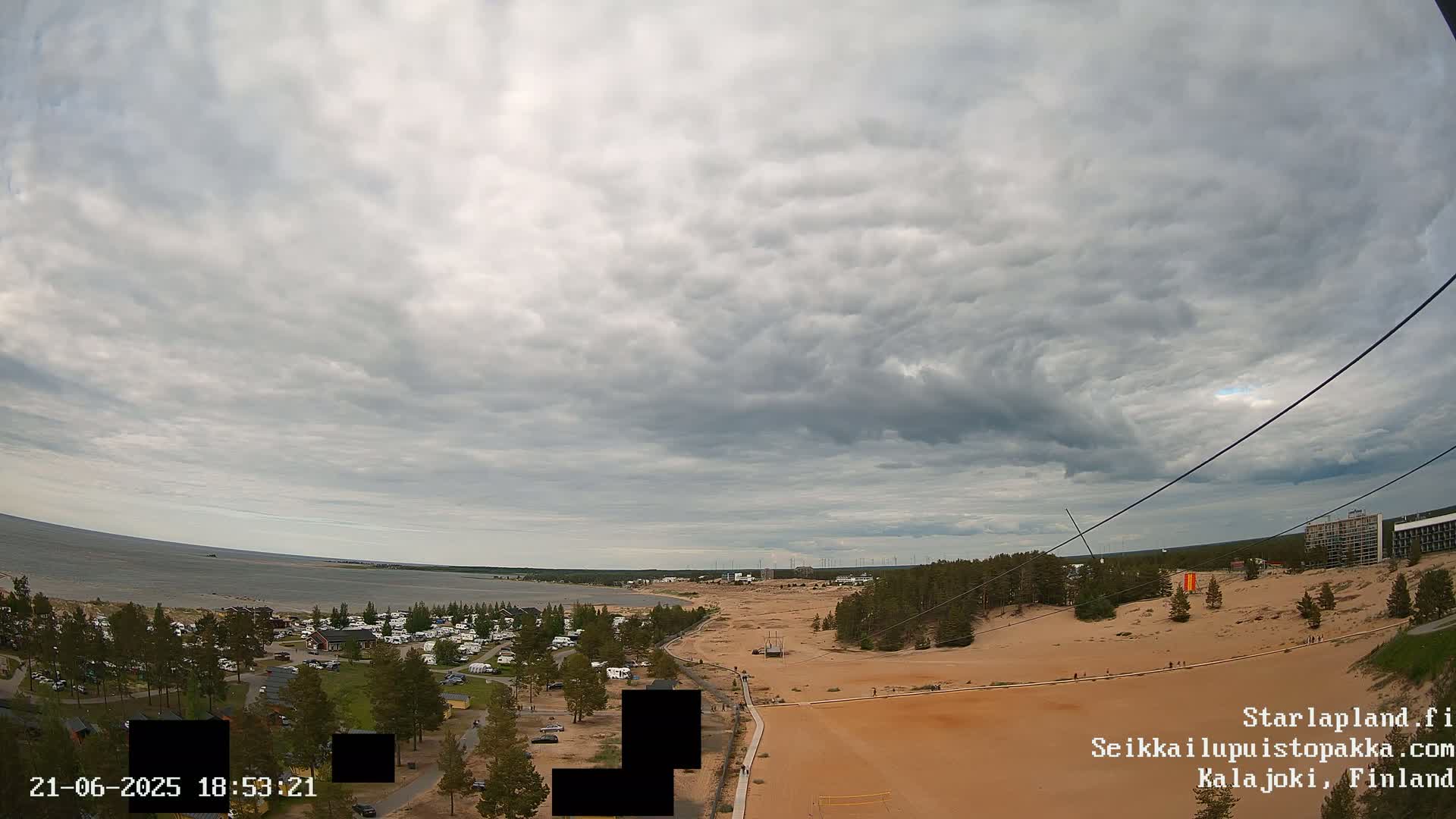 A high-angle view shows a beach, a sandy area, and a wooded area under a mostly cloudy sky.