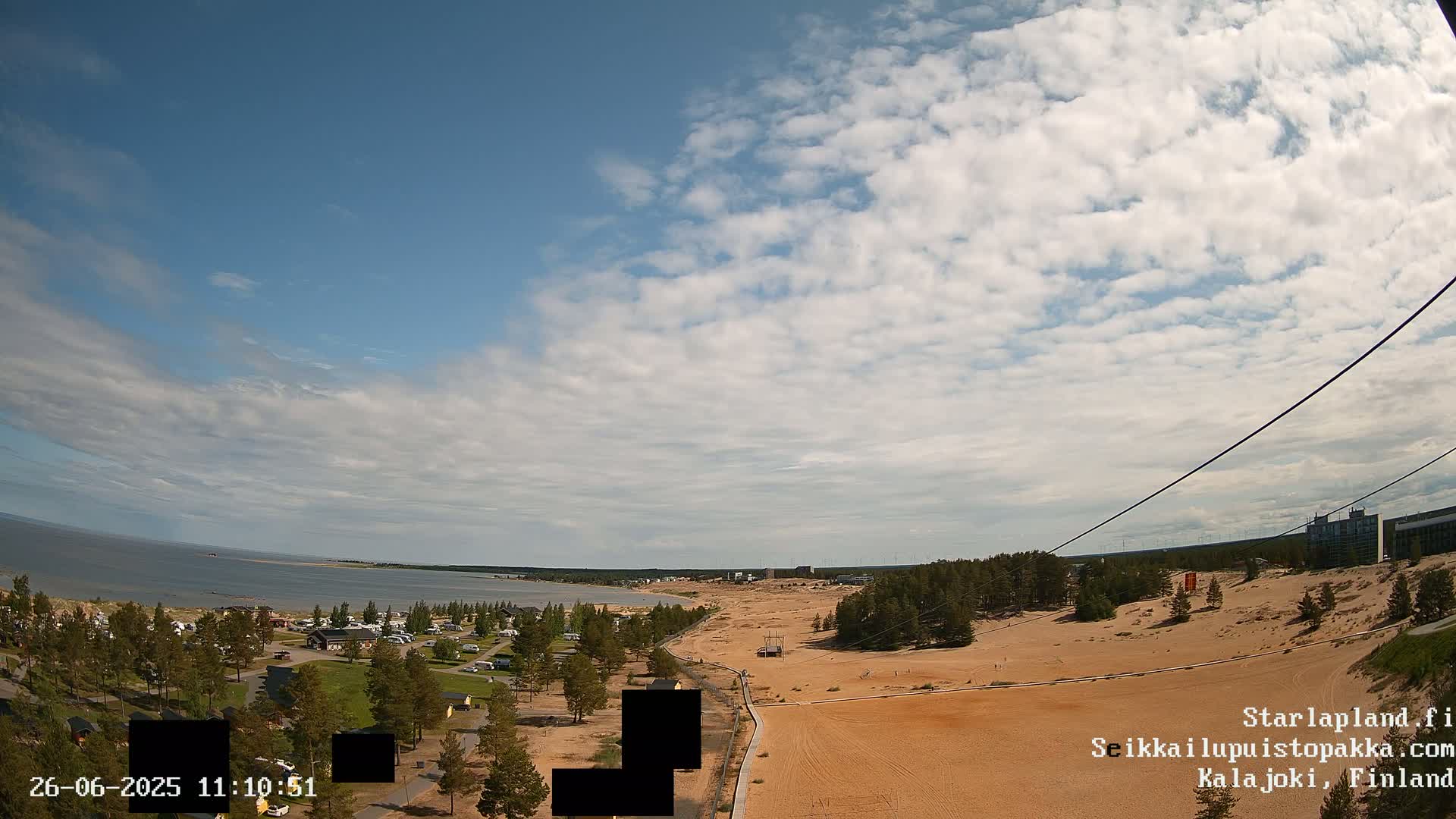 A mostly sunny day shows a sandy landscape next to a calm body of water, with a small town and forest visible in the distance.