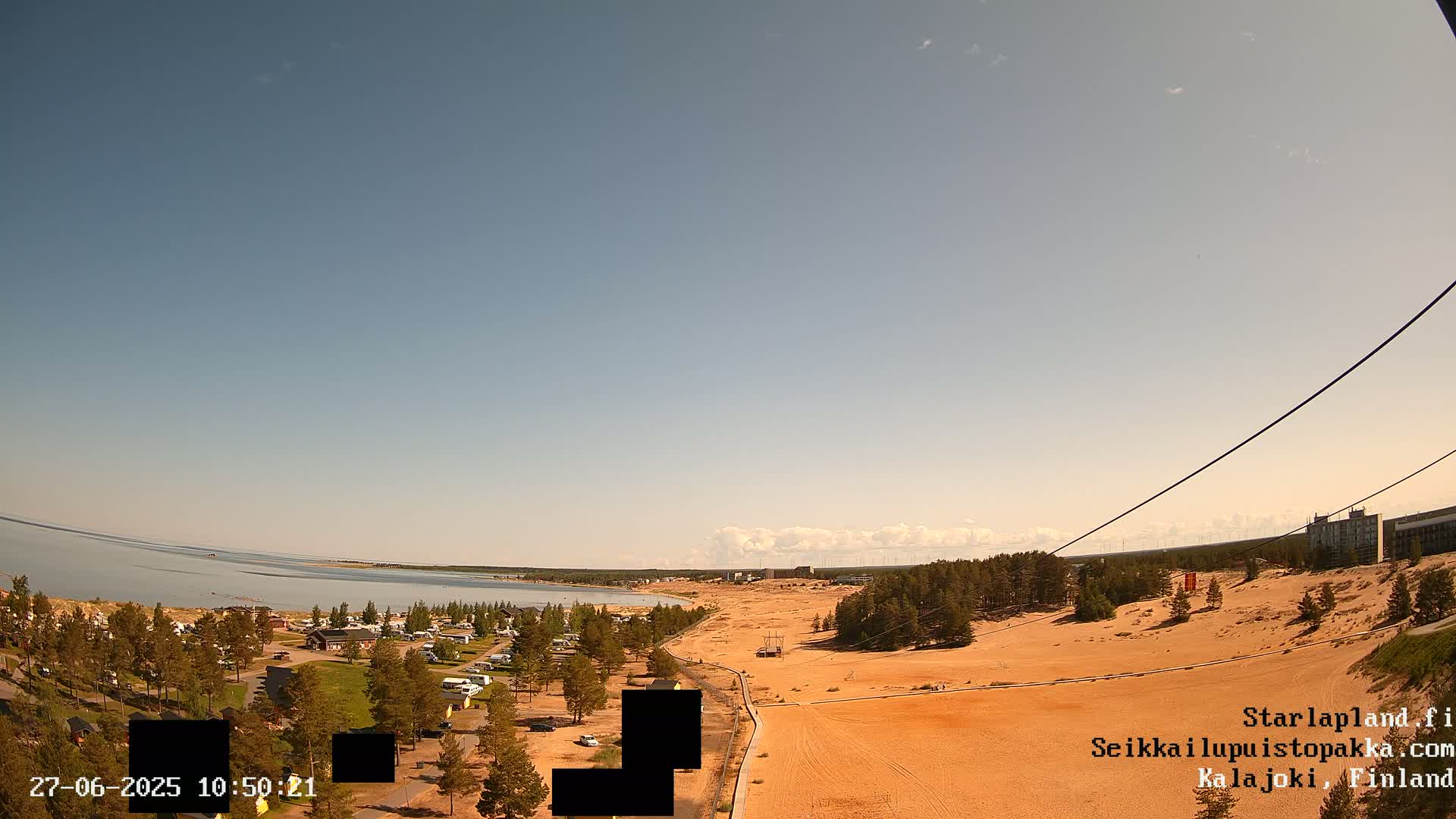 A mostly clear sky overlooks a sandy landscape next to a calm body of water, with a campground and some buildings visible in the distance.