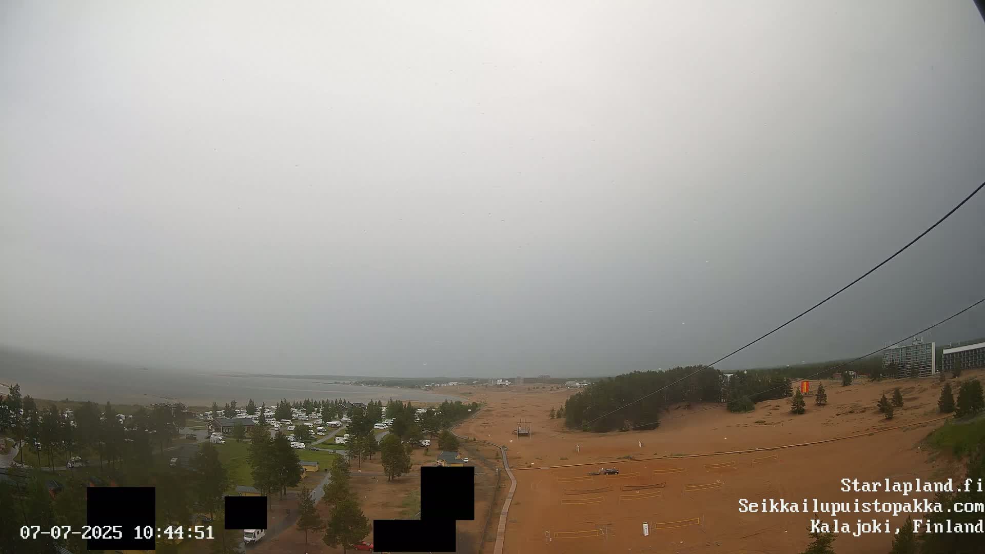 A high-angle, long shot shows a sandy area with a beach and calm body of water in the background under an overcast sky.