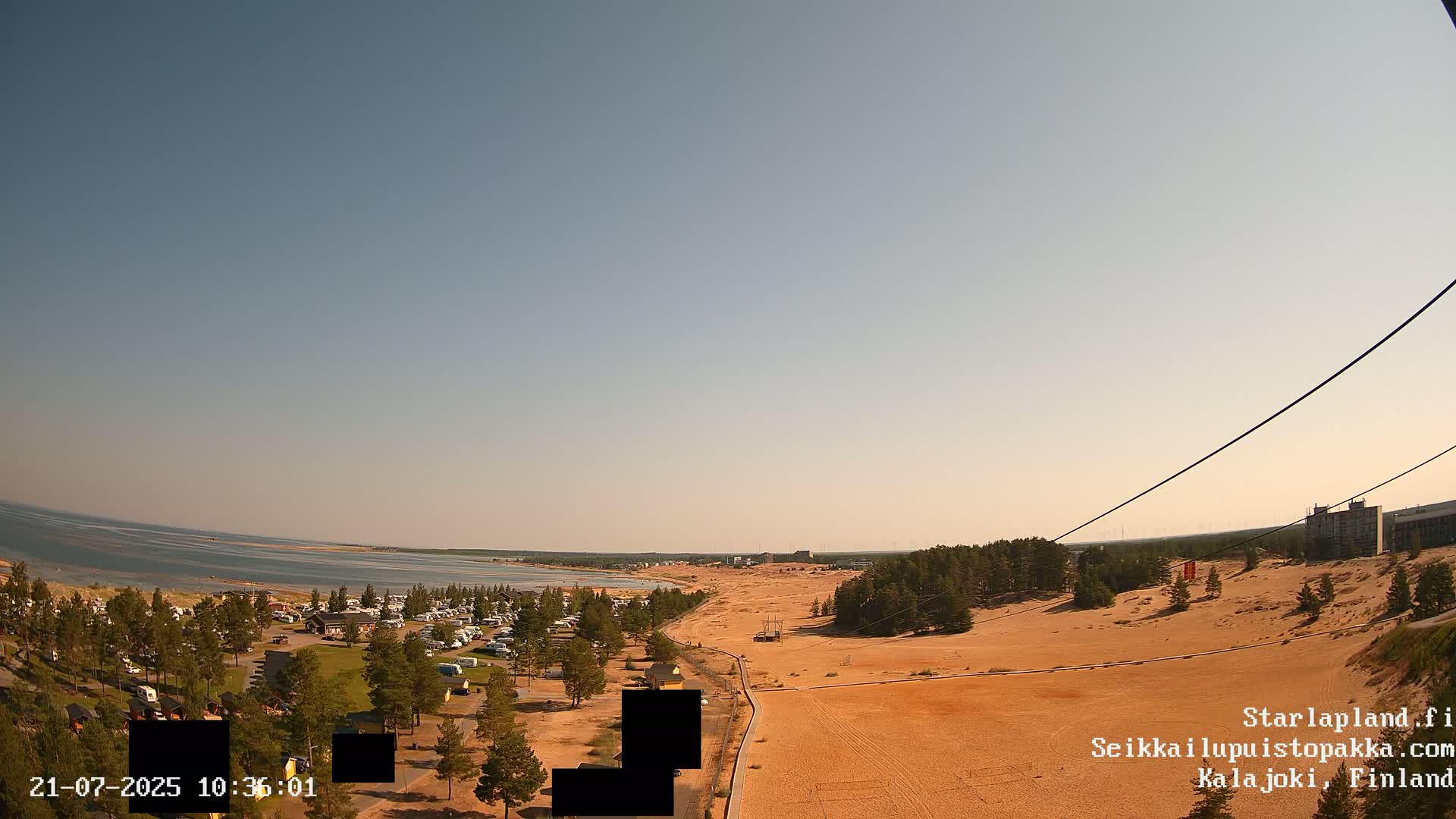 A high-angle view shows a sandy landscape with a campsite, a body of water, and a clear sky under bright sunlight.