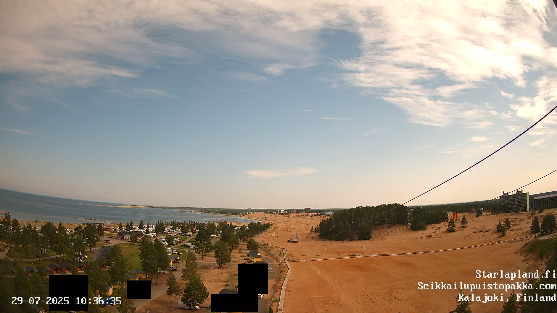 An elevated view shows a sandy landscape with a coastline and calm blue water under a partly cloudy sky.