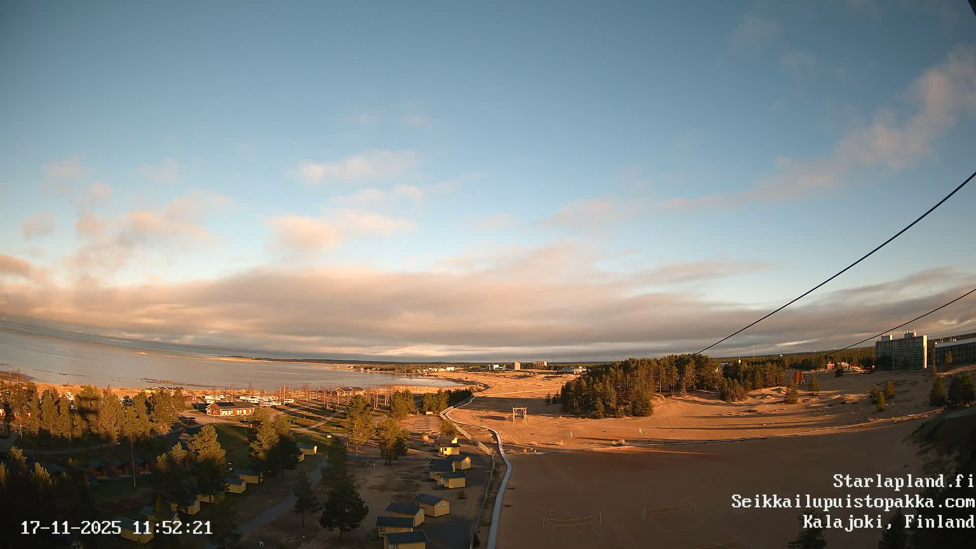 A wide outdoor shot captures a coastal landscape featuring a calm body of water, extensive sandy dunes, pine forests, small cabins, and distant modern buildings, all under a clear blue sky with scattered pinkish-orange clouds.