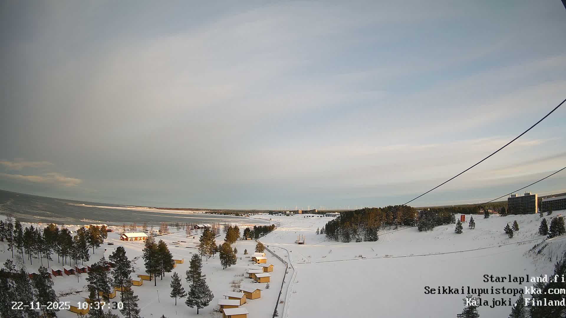 A wide, snow-covered winter landscape under an overcast sky showcases small yellow cabins among frosted evergreen trees, extending to a partially frozen body of water and a distant horizon dotted with buildings and wind turbines.