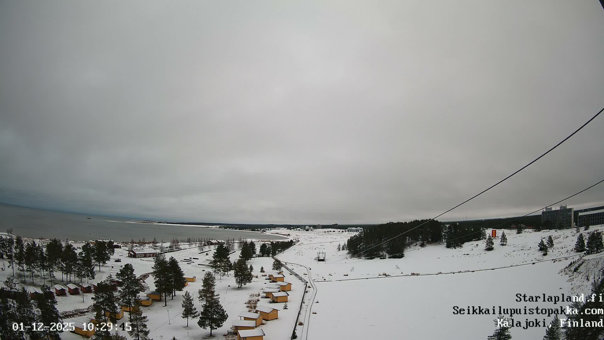 A snow-covered winter landscape, featuring a large body of water partially frozen on the left, numerous small cabins and pine trees, a winding outdoor track, and modern buildings in the distance, is visible under a uniformly overcast sky.