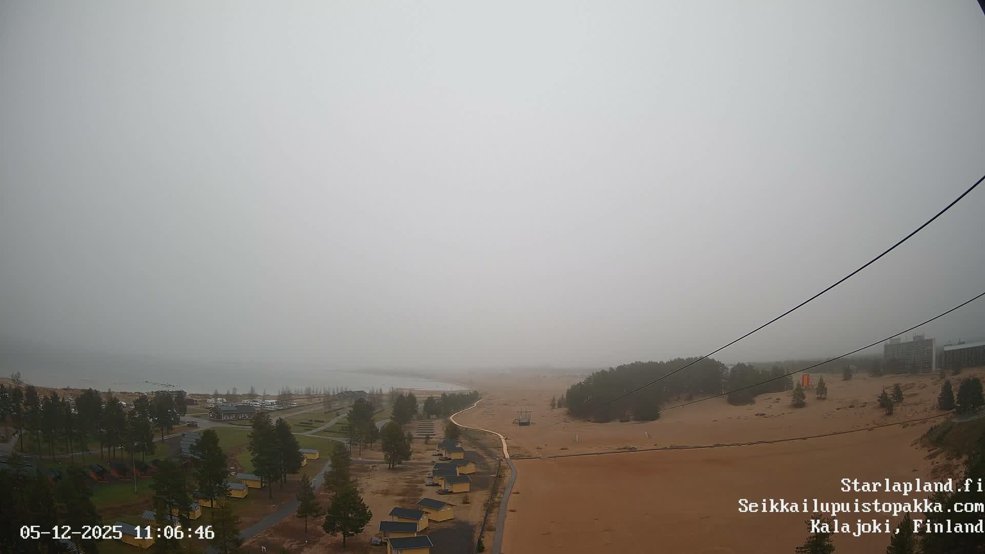 A wide, sandy landscape with scattered pine trees and yellow cabins borders a calm body of water on a heavily overcast and gloomy day.