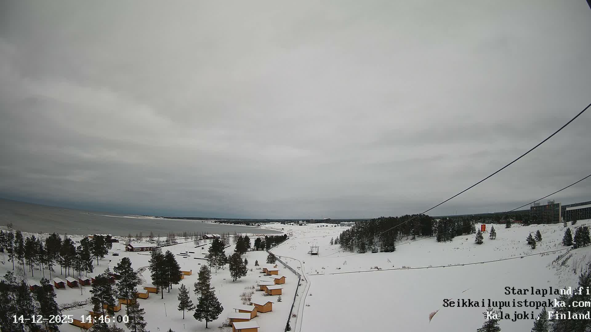 A wide, sandy landscape with scattered pine trees and yellow cabins borders a calm body of water on a heavily overcast and gloomy day.