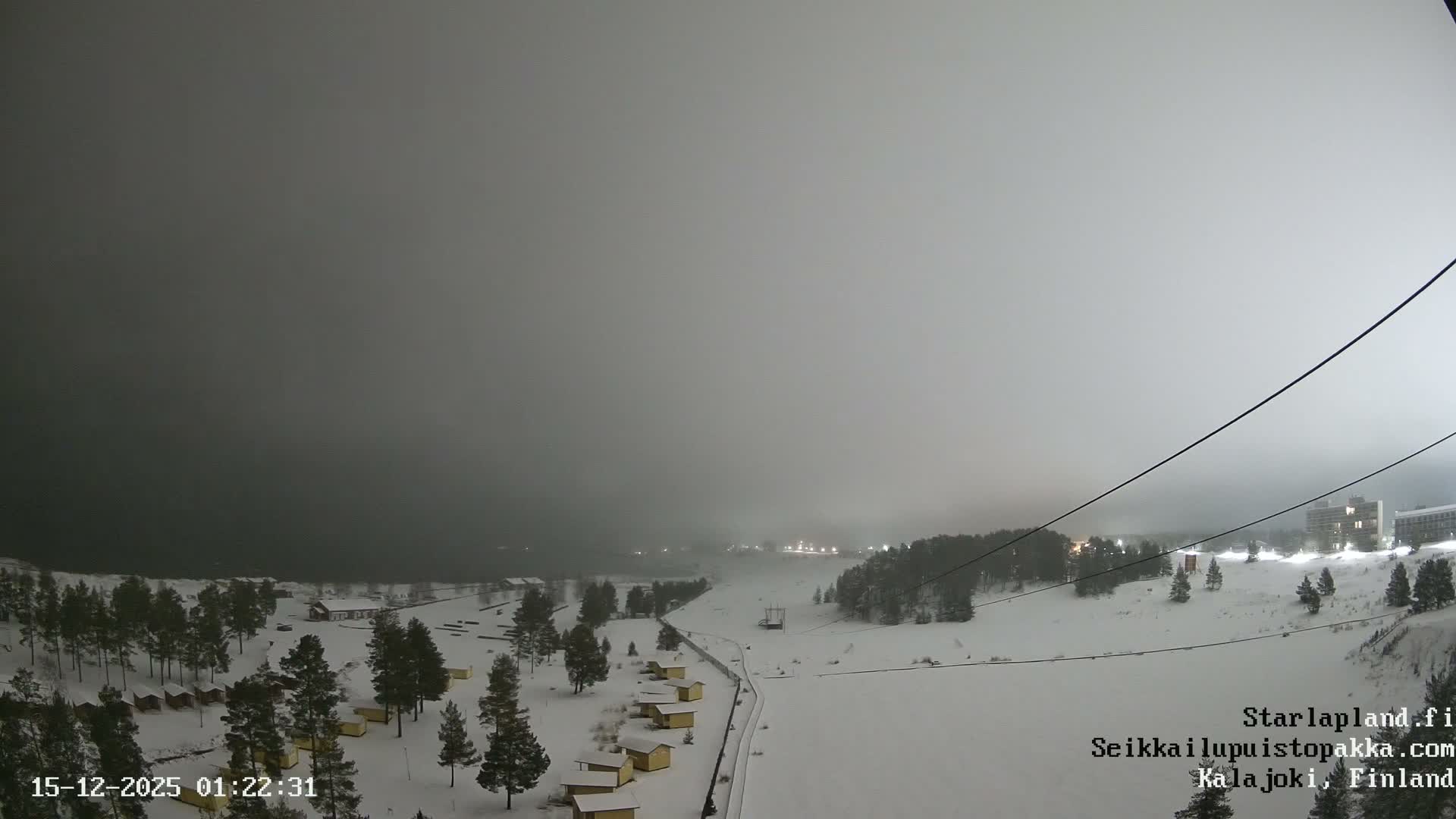 A wide, sandy landscape with scattered pine trees and yellow cabins borders a calm body of water on a heavily overcast and gloomy day.