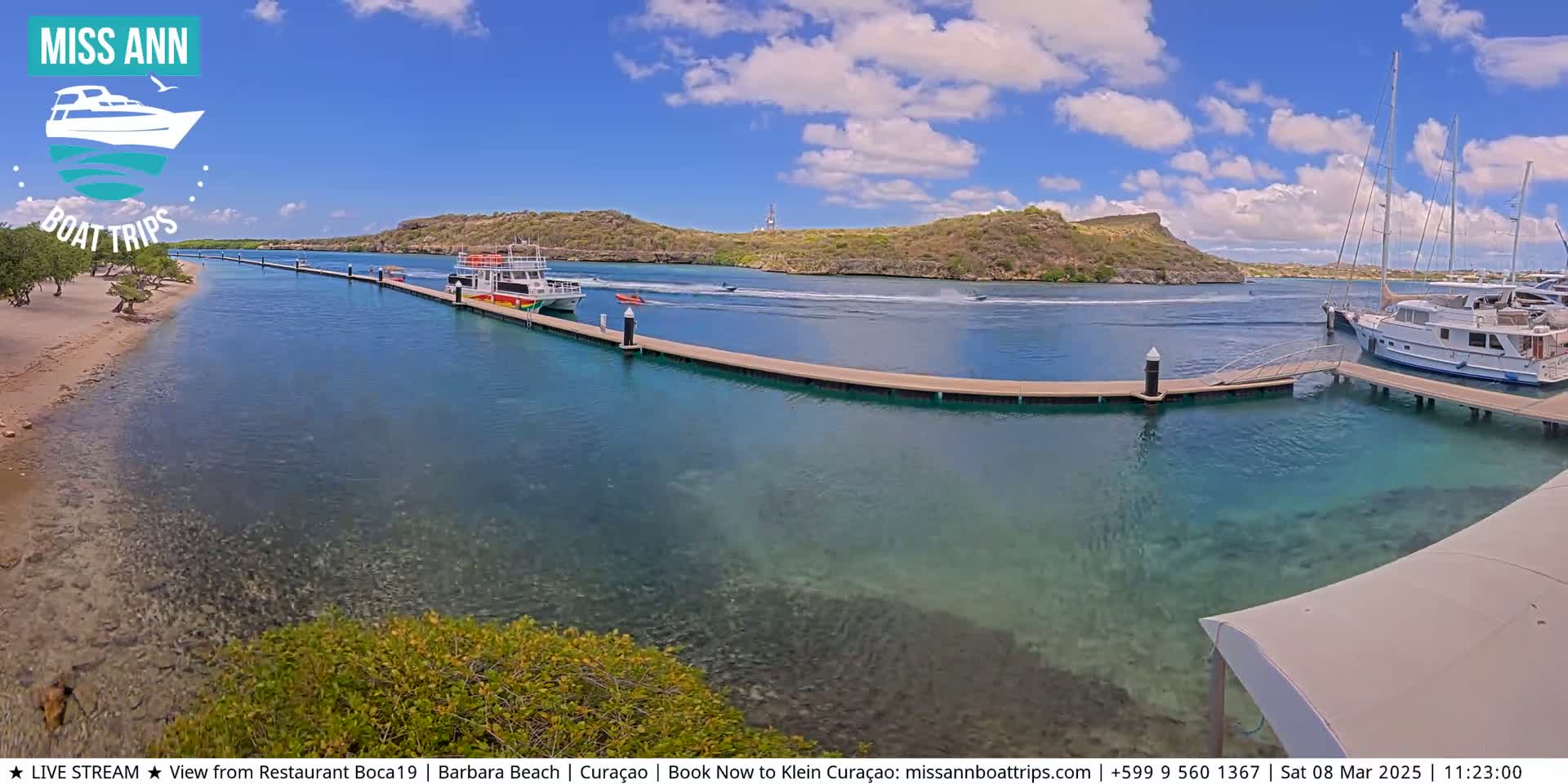 A sunny day shows a calm, clear turquoise body of water with several boats docked at a pier near a sandy beach and a hilly, green shoreline.