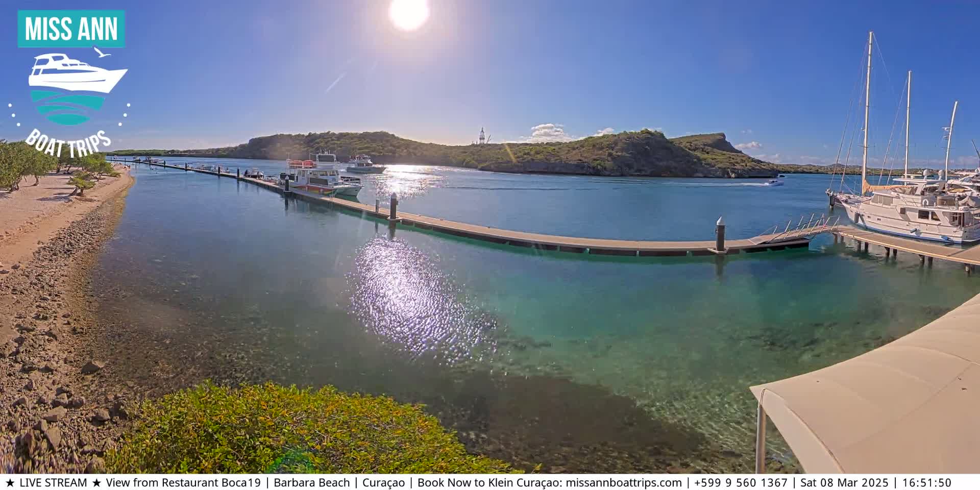 A sunny day shows clear, shallow turquoise water in a marina with several boats docked, a sandy beach, and rocky hills in the background.