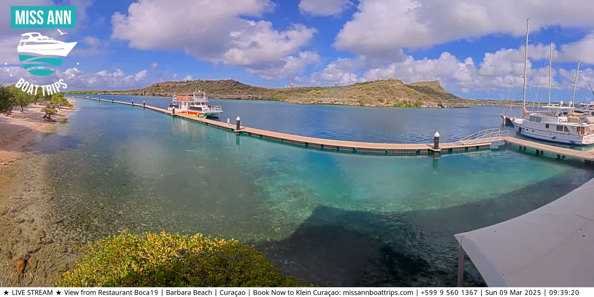 A sunny day reveals a calm, clear turquoise body of water with docked boats near a sandy shore and low-lying hills in the background.