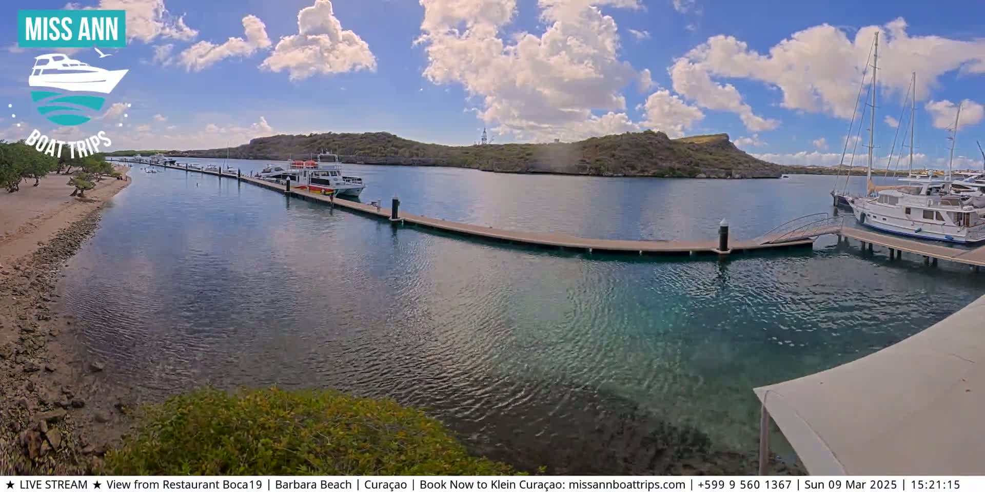 A sunny day shows a calm harbor with several boats docked at a long pier,  a sandy beach, and low-lying hills in the background.