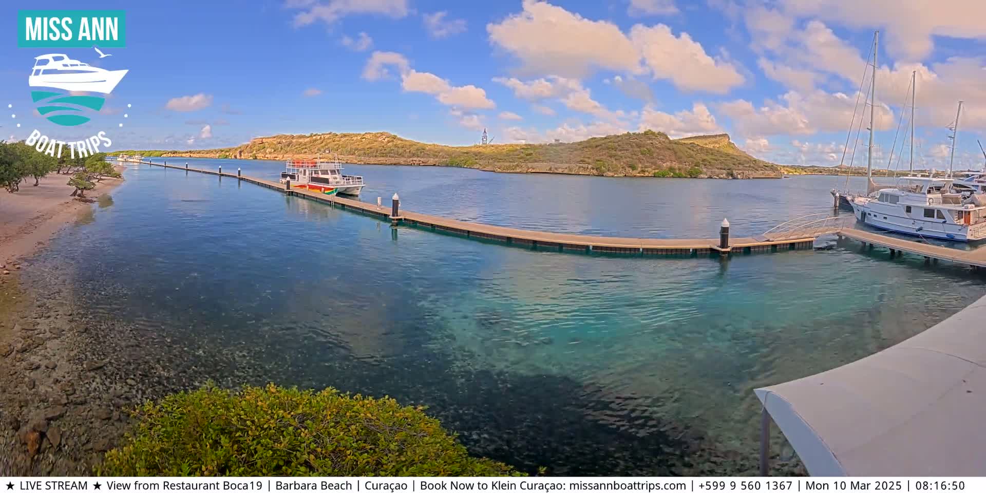 A calm, sunny day shows a marina with several boats docked, clear turquoise water, and a sandy beach with vegetation in the background.