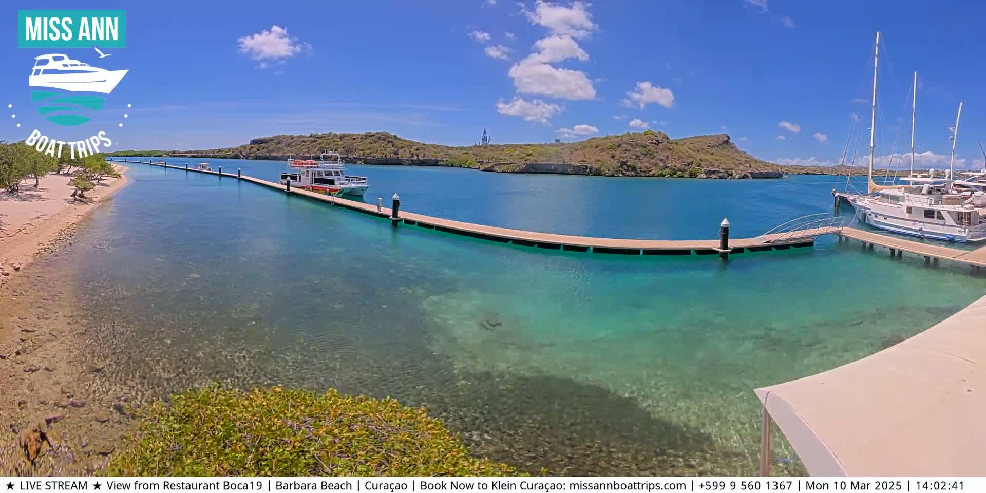 A clear, sunny day reveals a calm, turquoise bay with boats docked at a long pier, near a sandy beach and low-lying hills.