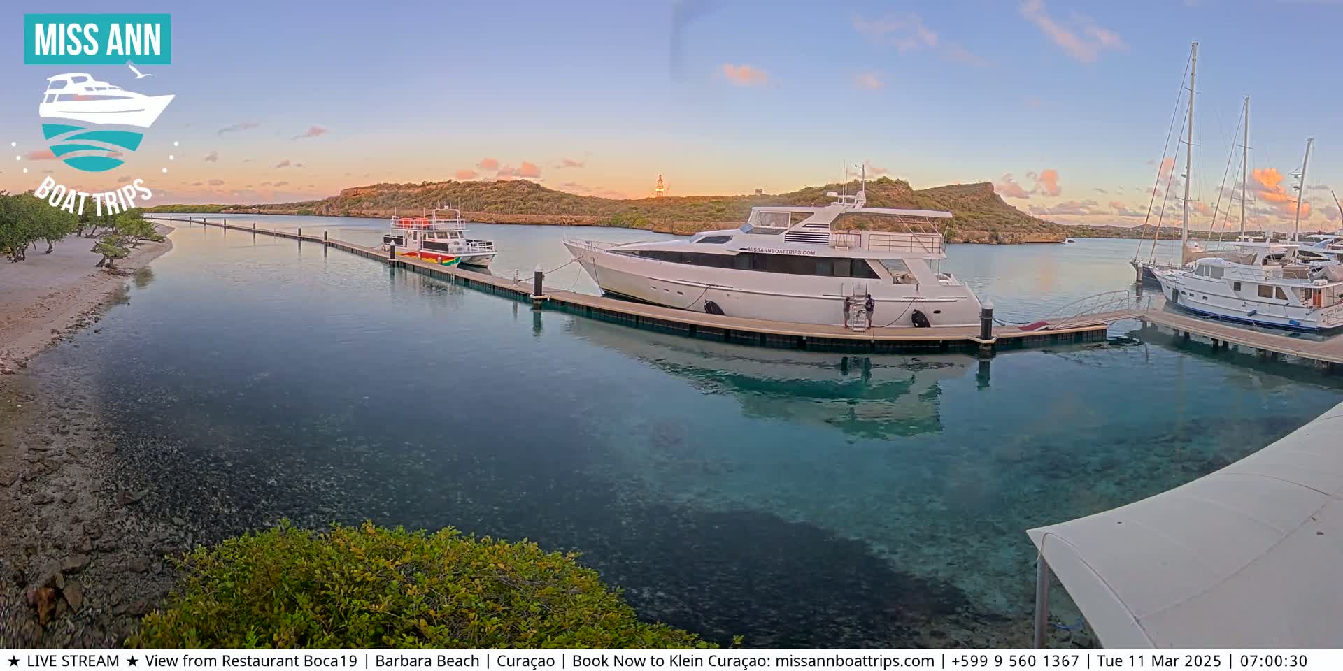 Several boats are docked at a marina in calm, clear water under a sunset sky.