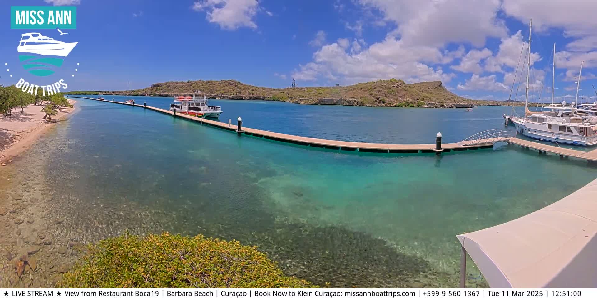 A sunny day shows a turquoise bay with several boats docked at a long pier extending from a sandy shore, backed by low-lying hills.