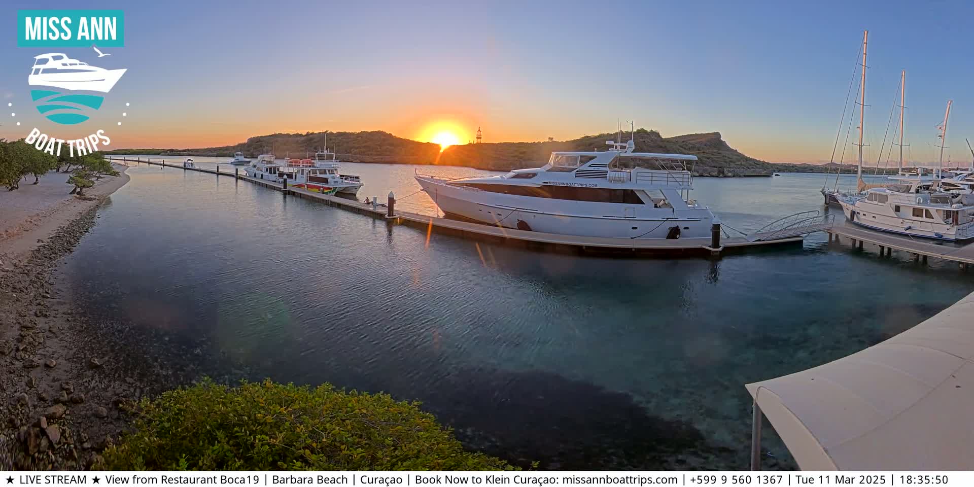 Several boats are docked at a marina during a sunset, with calm water and a sandy shore visible in the foreground.