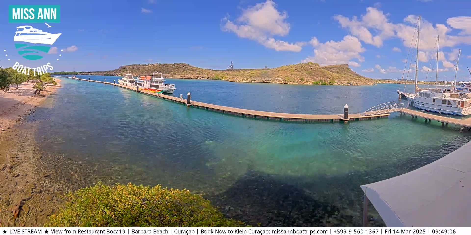 A sunny day shows a calm, clear turquoise body of water with several boats docked at piers near a sandy shore and a rocky, hilly island in the background.