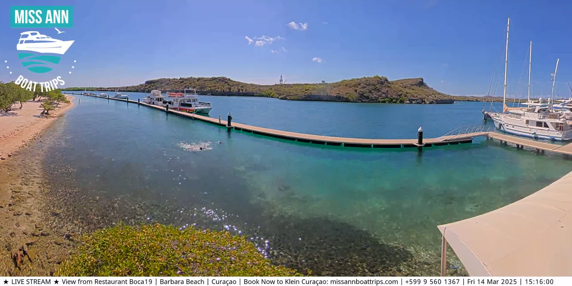 A calm, clear turquoise body of water with several boats docked at a pier, under a sunny sky.