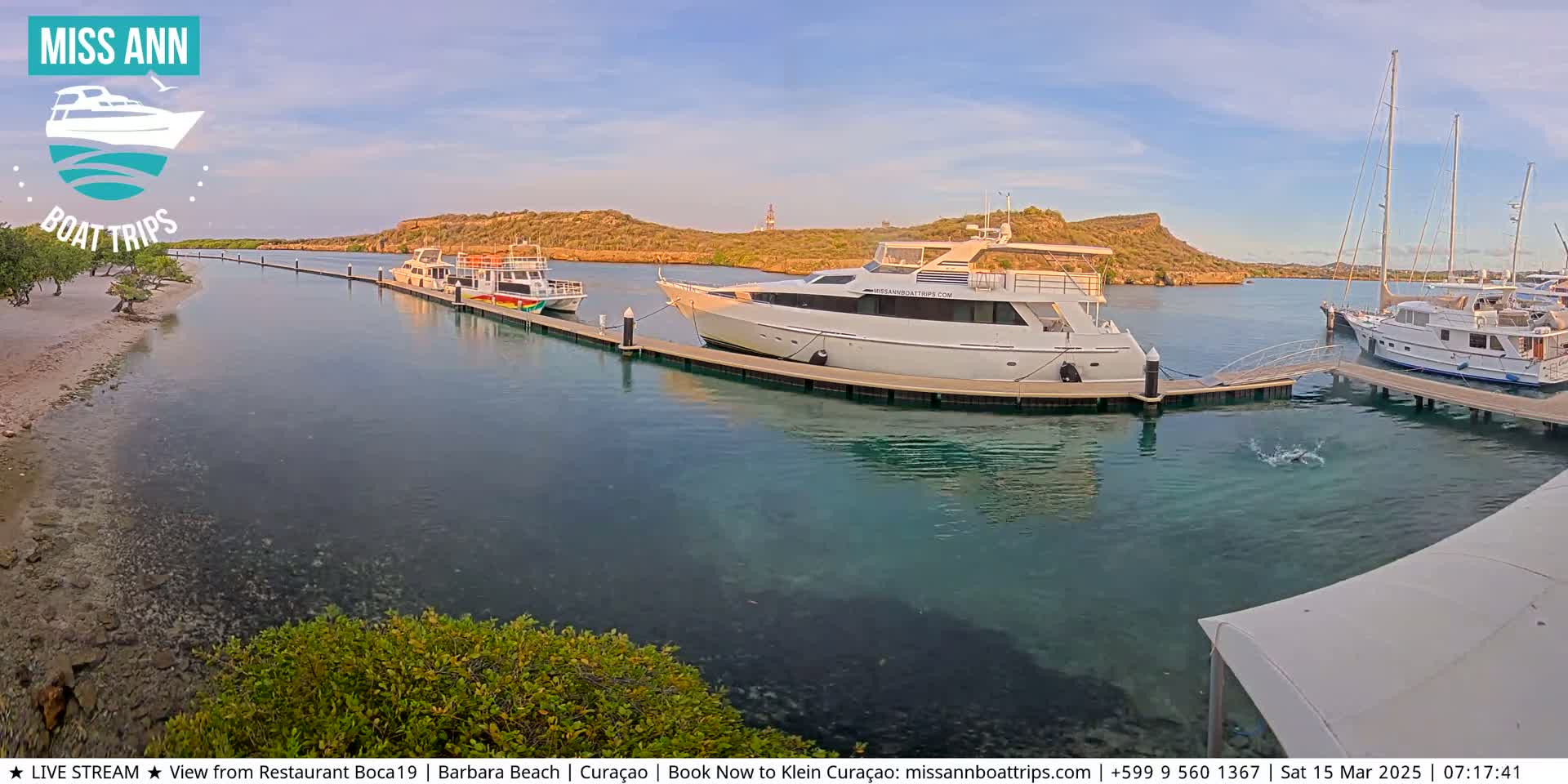 Several boats are docked at a marina on calm, clear water next to a sandy shore and arid hills under a partly cloudy sky.