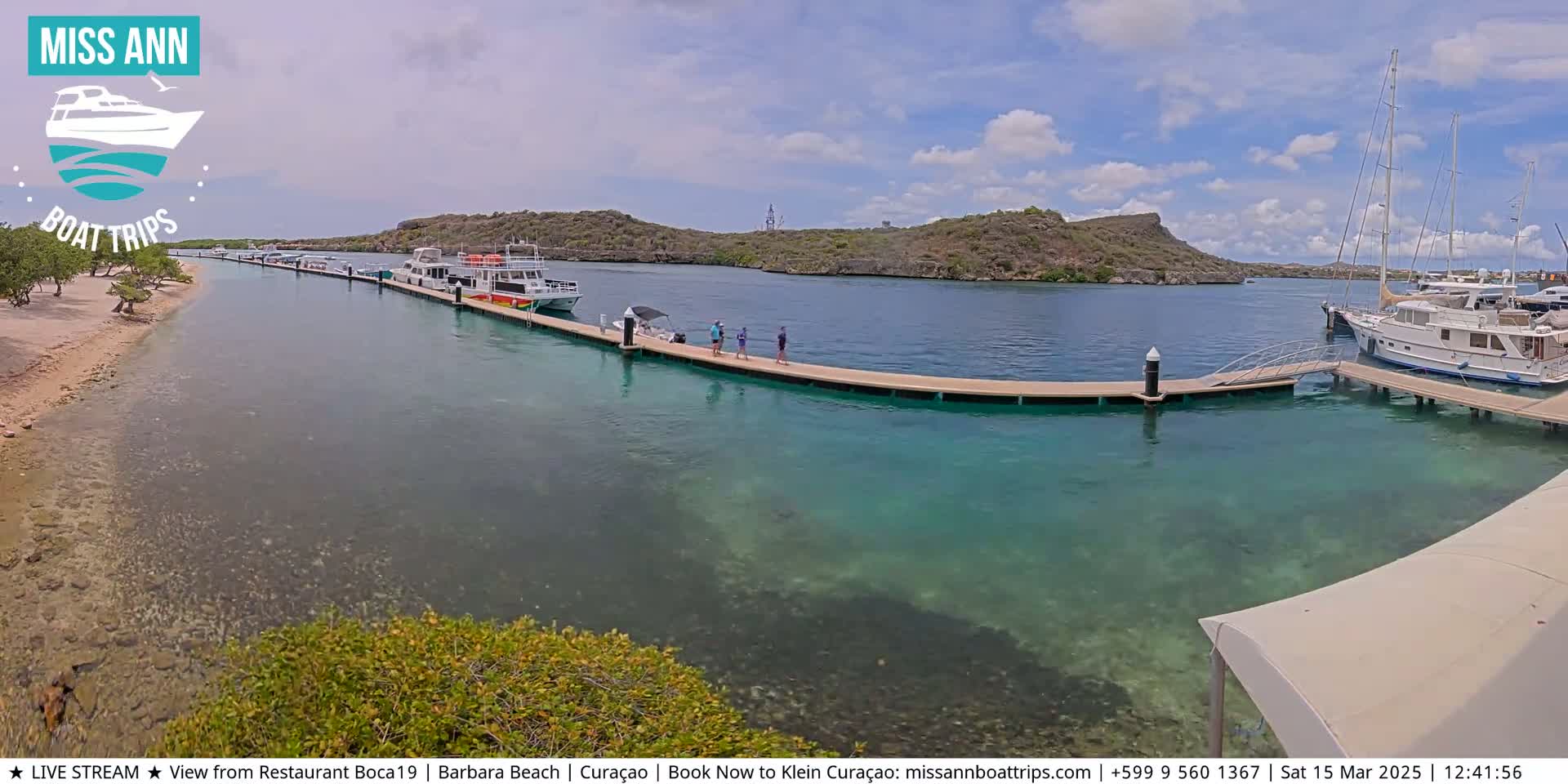 A calm, sunny day shows a marina with several boats docked, clear turquoise water, and people standing on a pier next to a sandy shore with low vegetation.