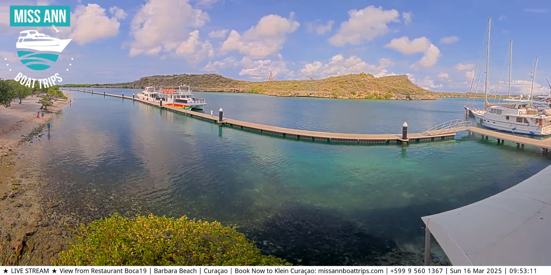 A sunny day reveals a calm bay with several boats docked at piers, a sandy beach with people on it, and a verdant hillside in the background.