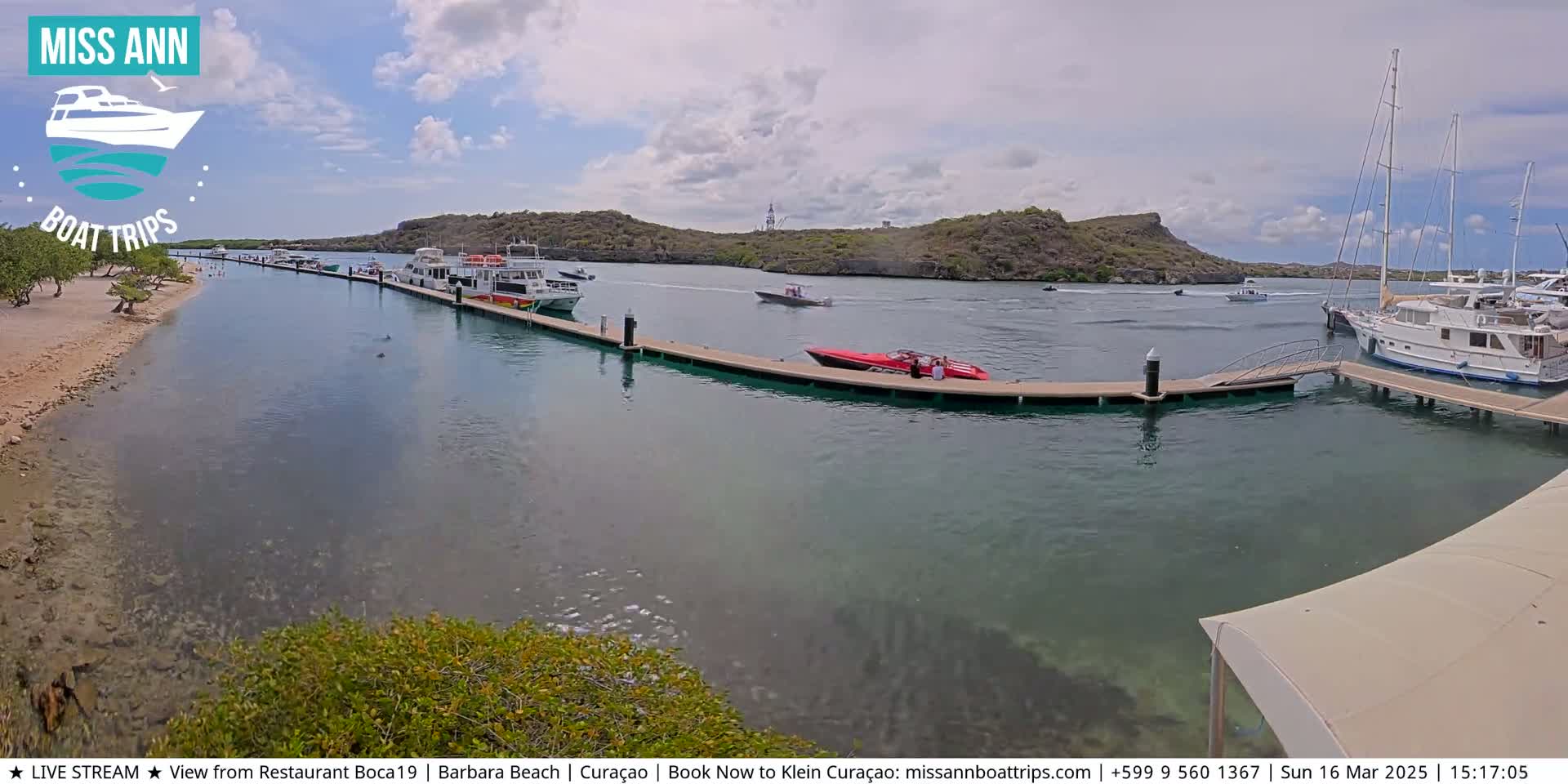 A calm, sunny day shows several boats docked at a marina, with additional boats and personal watercraft in the clear water near a sandy beach and rocky hills.