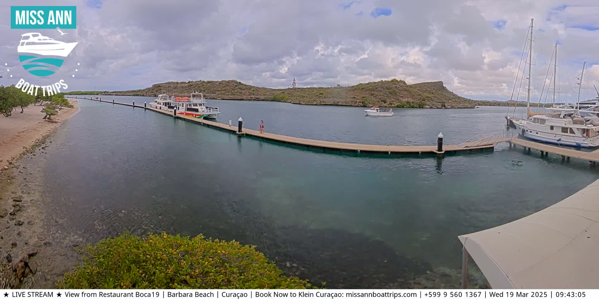 Several boats are docked at a marina on a calm, partly cloudy day, with a sandy beach and green hills visible in the background.