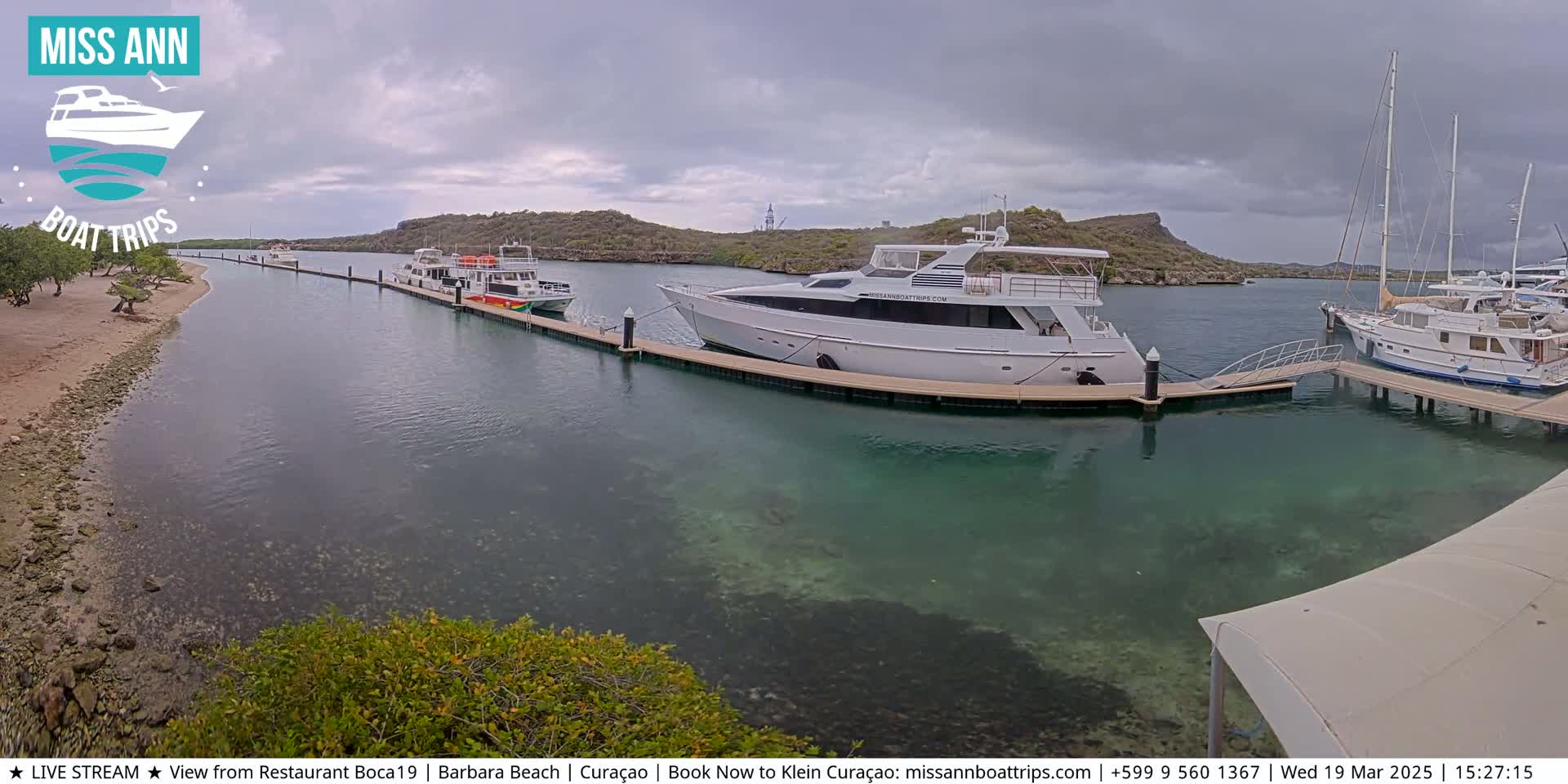 Several boats are docked at a marina on a partly cloudy day, with clear, shallow water visible near the docks.