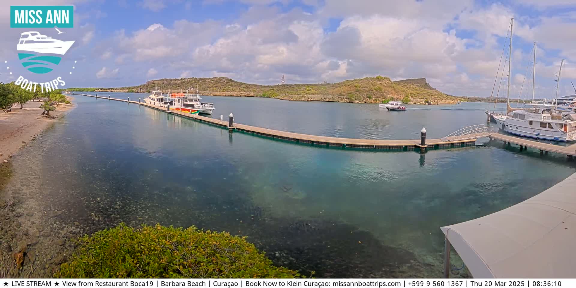 A calm, sunny day shows several boats docked at a marina next to a sandy beach and a hilly, arid landscape.