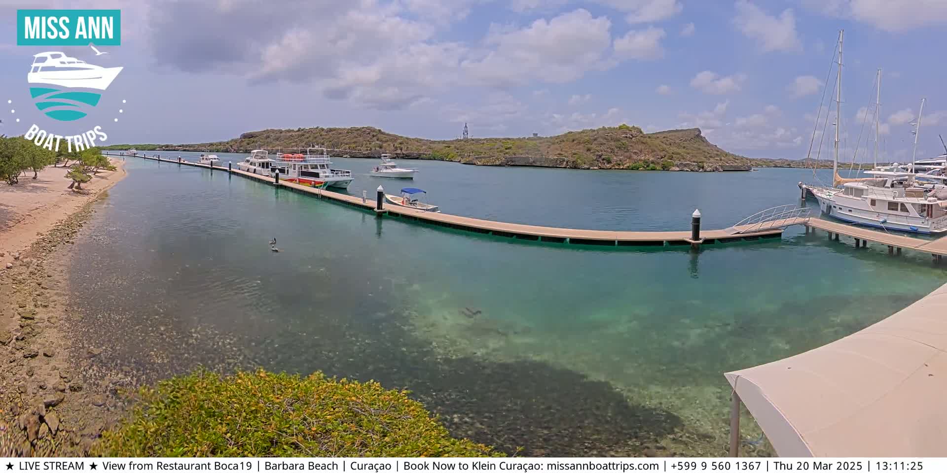 Several boats are moored at a dock in clear, shallow turquoise water near a sandy shore and low-lying, rocky hills under a partly cloudy sky.