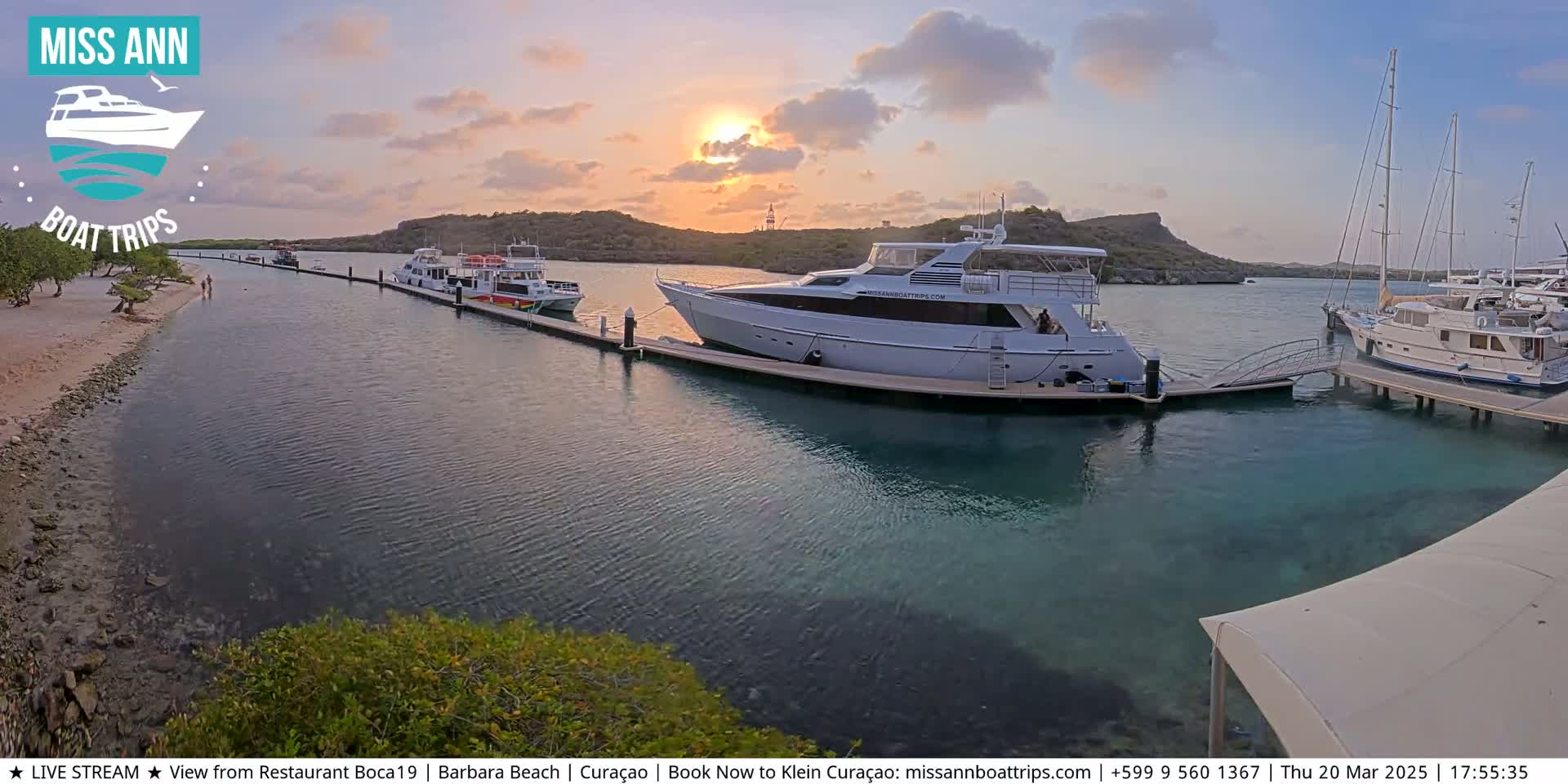 Several boats are docked at a marina during sunset, with calm water and a sandy shore visible in the foreground.