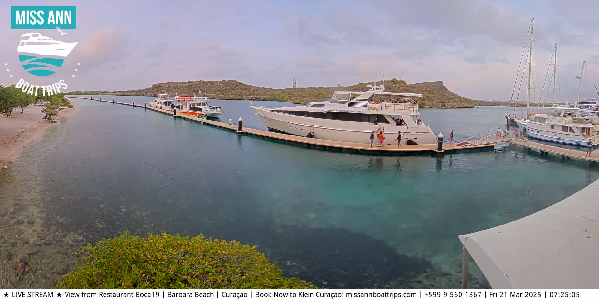 Several boats are docked at a marina on a partly cloudy day, with clear turquoise water visible around them.