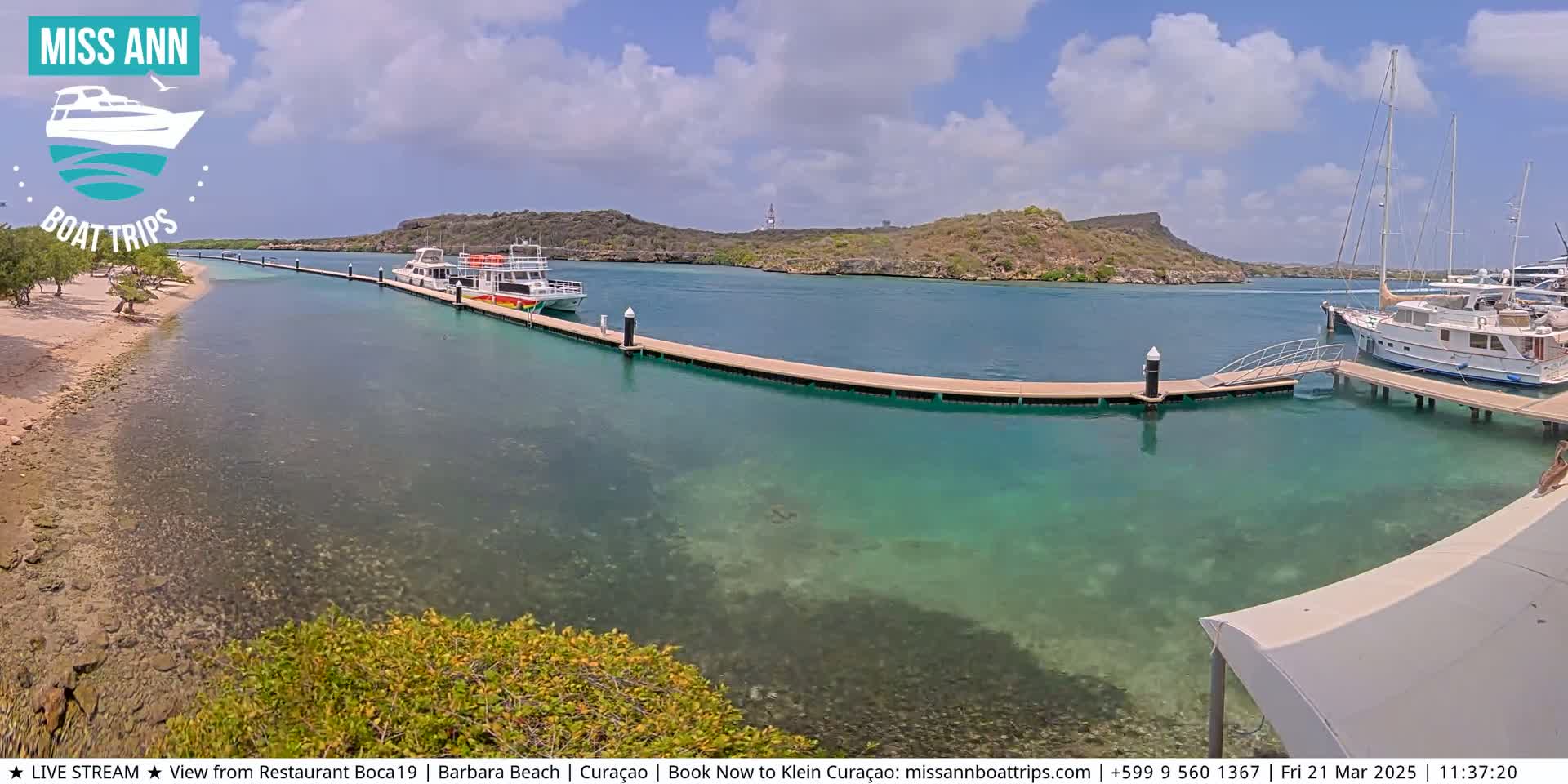 A calm, sunny day reveals a clear turquoise harbor with several boats docked at a long pier, near a sandy beach and small, rocky islands.