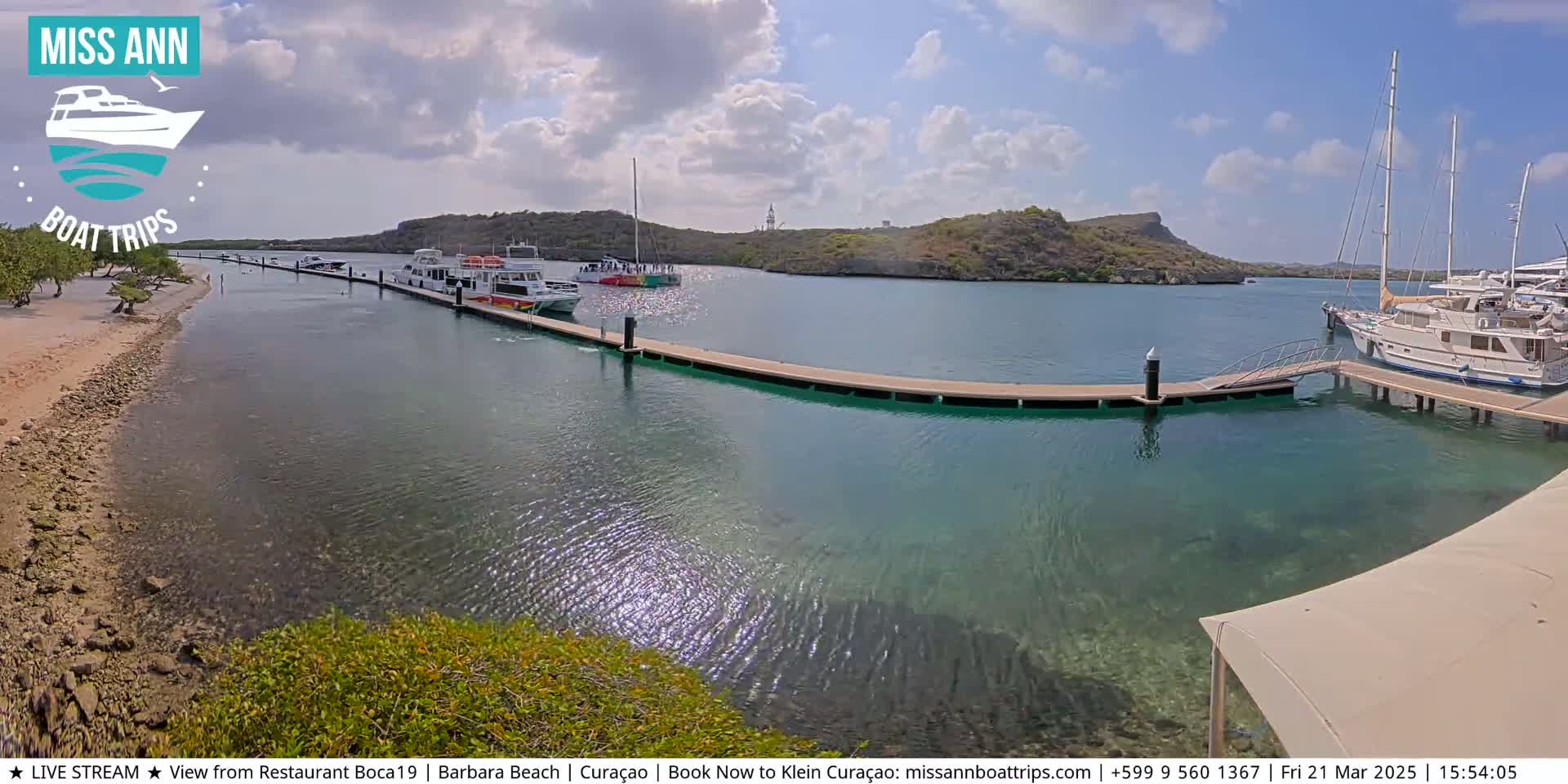 Several boats are docked at a marina on a partly cloudy day, with clear, shallow turquoise water visible around the docks and a sandy beach in the foreground.