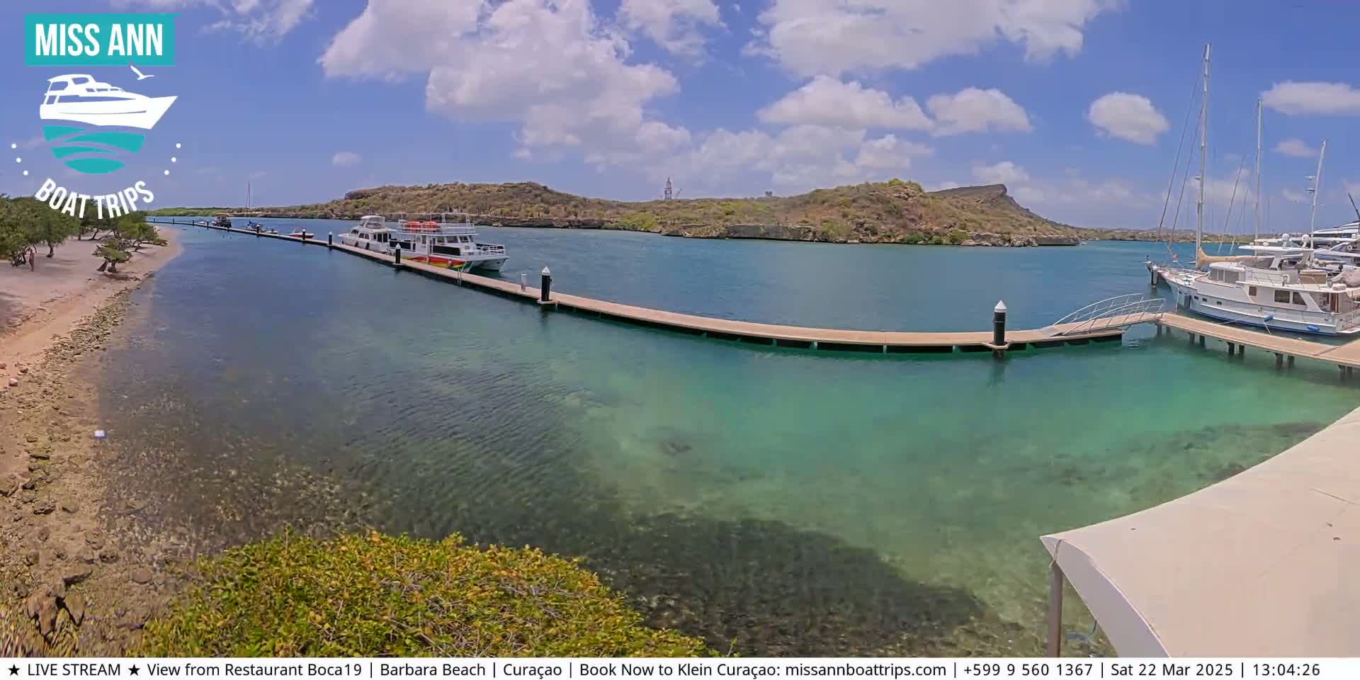 A tranquil marina with clear, shallow turquoise water features several boats docked at a long wooden pier, with a sandy beach and low-lying vegetation visible in the foreground under a partly cloudy sky.