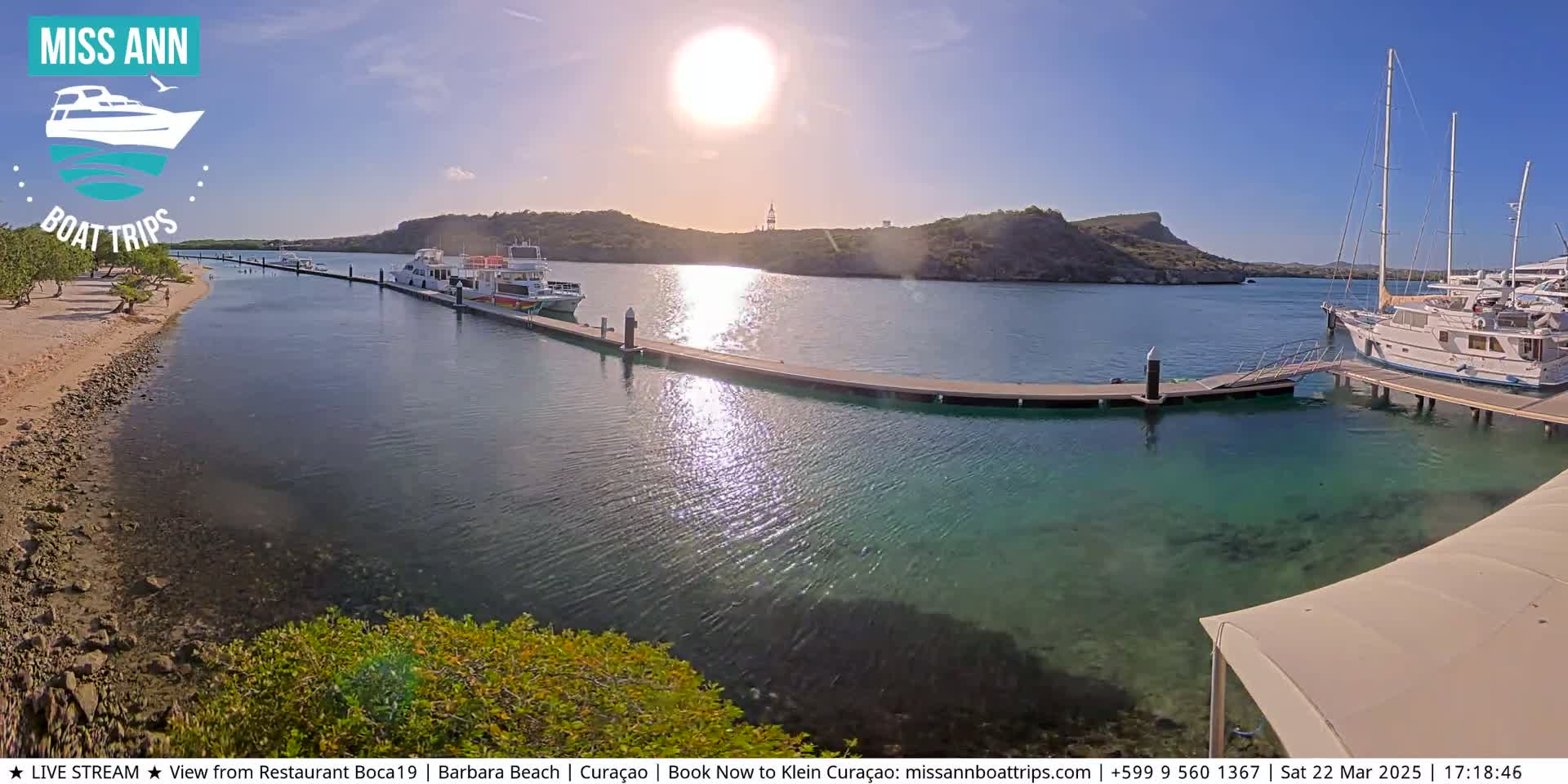 Several boats are docked at a marina on a sunny day, with clear, shallow turquoise water visible around the docks and a sandy beach nearby.