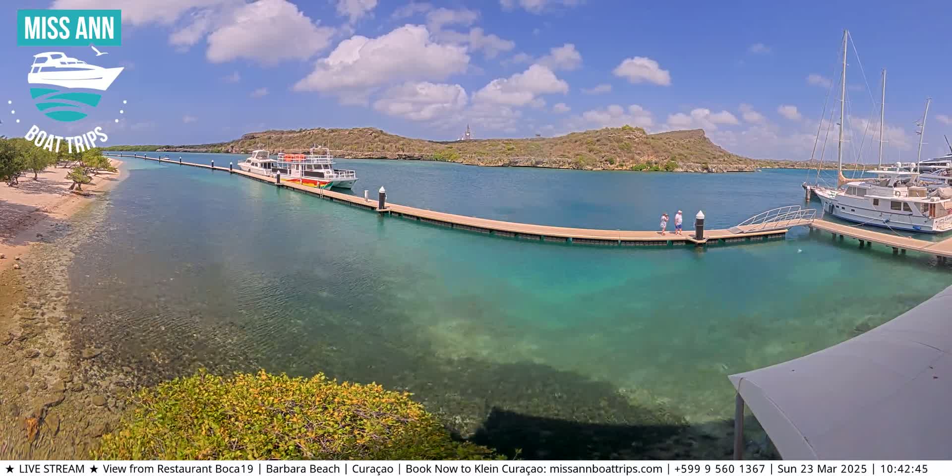 A sunny day shows a clear, turquoise harbor with boats docked at a long pier, and two people walking on the pier.