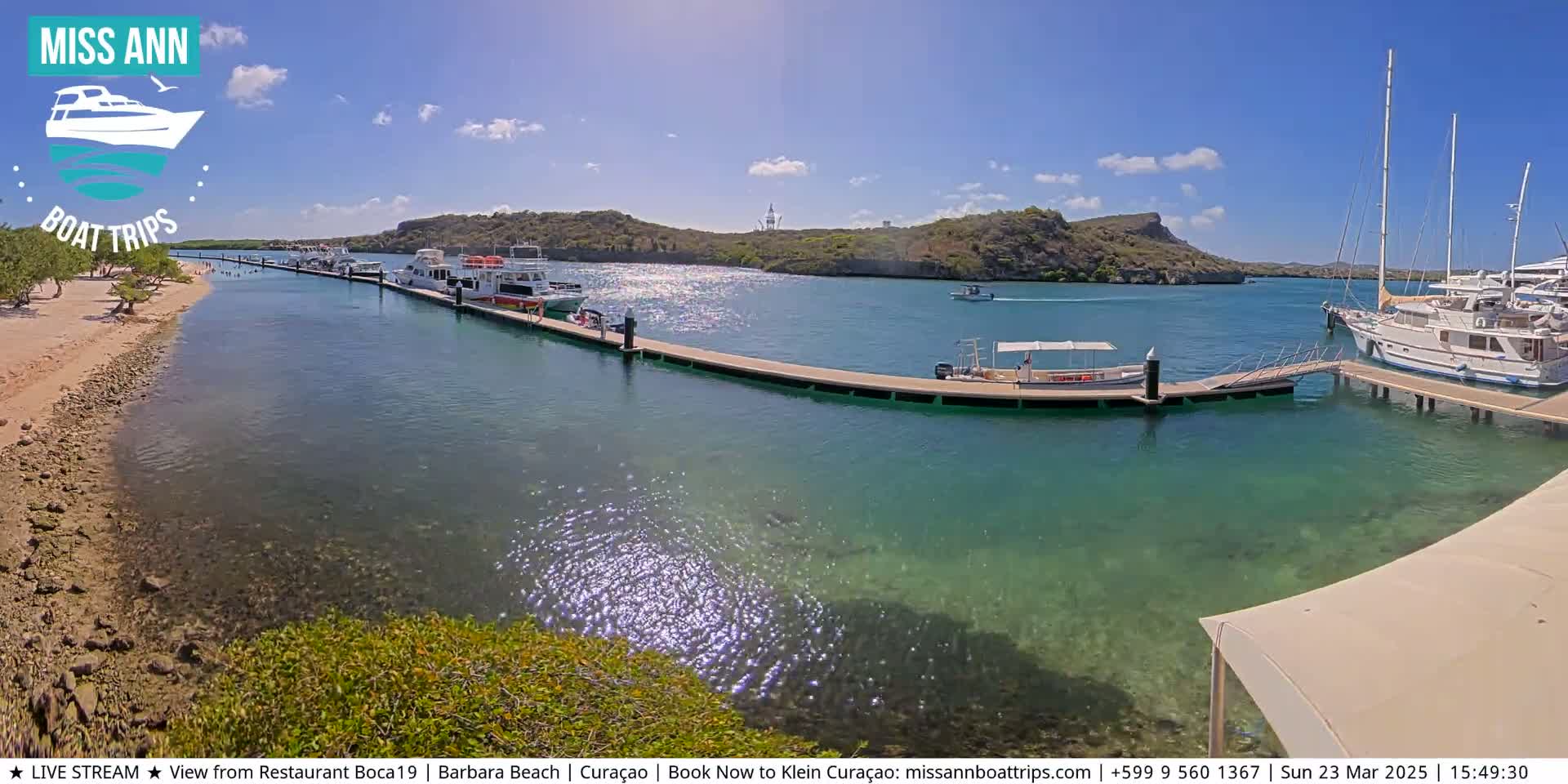 A sunny day reveals a calm, clear bay with several boats docked at a pier, extending from a sandy shoreline toward hills under a partly cloudy sky.