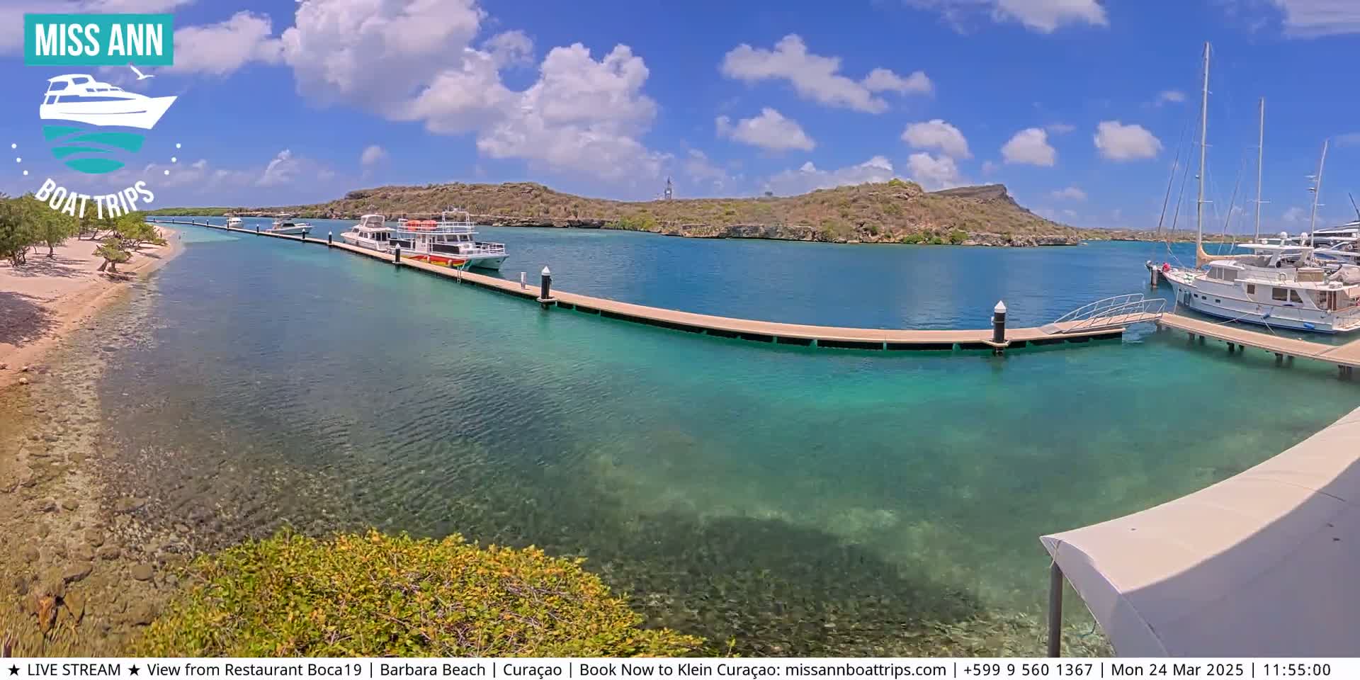 A sunny day reveals several boats docked at a marina with clear, turquoise water and a sandy beach in the background.