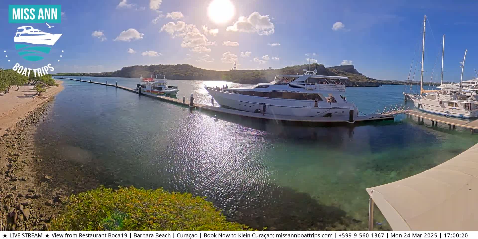 Several boats are docked at a marina on a sunny day with clear skies,  a sandy beach is visible in the foreground.