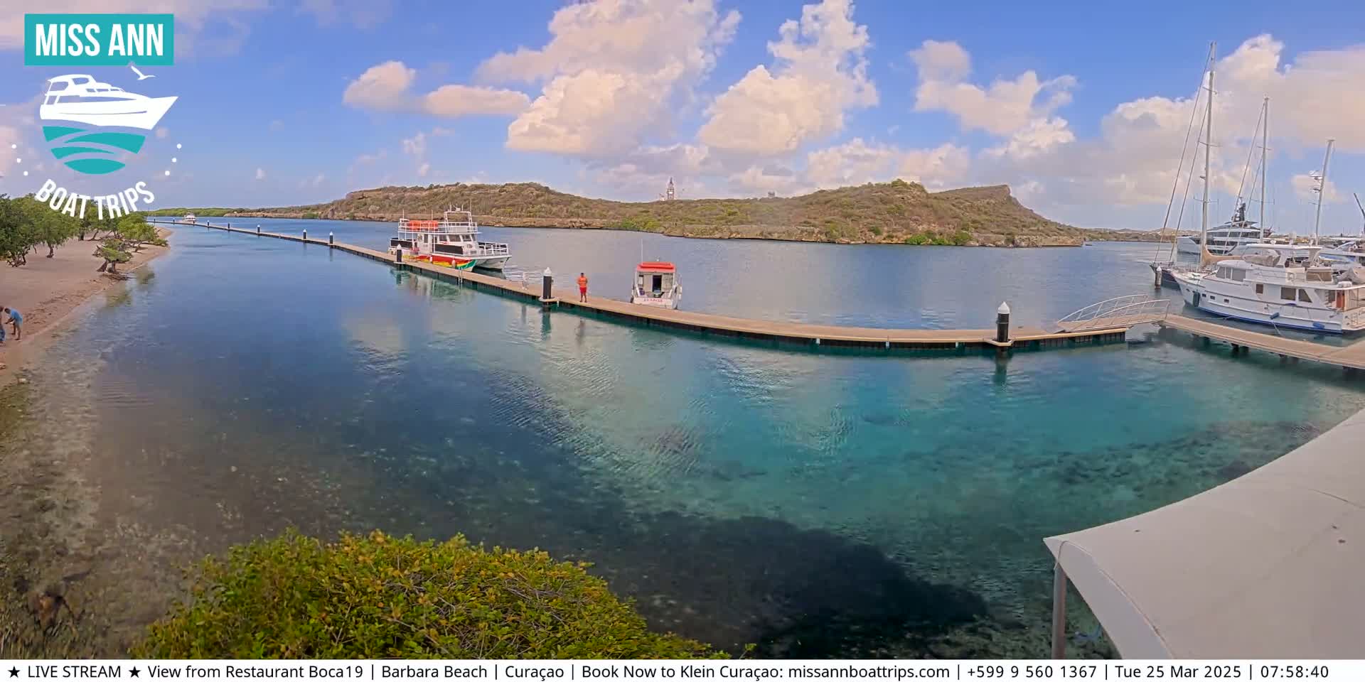 A calm, sunny day reveals a harbor with several boats docked at piers, clear turquoise water, and a sandy beach with sparse vegetation.