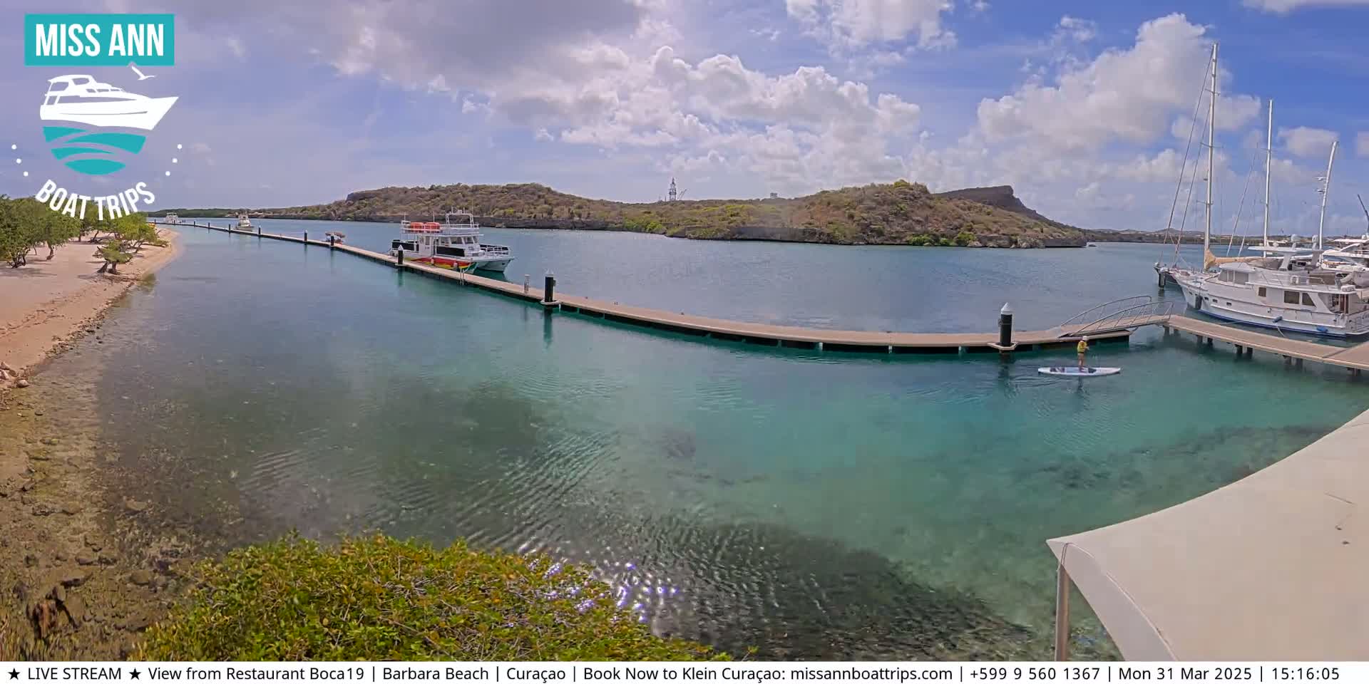 A calm, sunny day shows a marina with several boats docked, a person paddleboarding, and clear turquoise water.