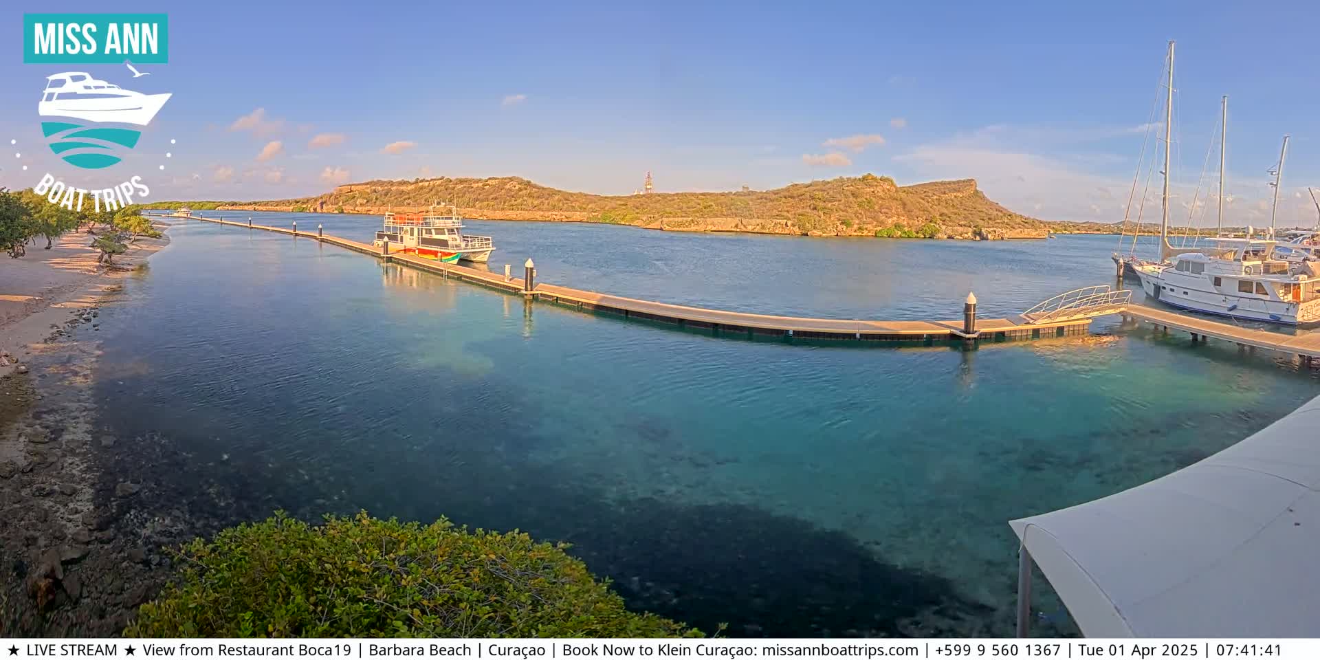 A calm, clear turquoise body of water shows boats docked at piers near a sandy shoreline and a hilly, arid landscape under a sunny sky.