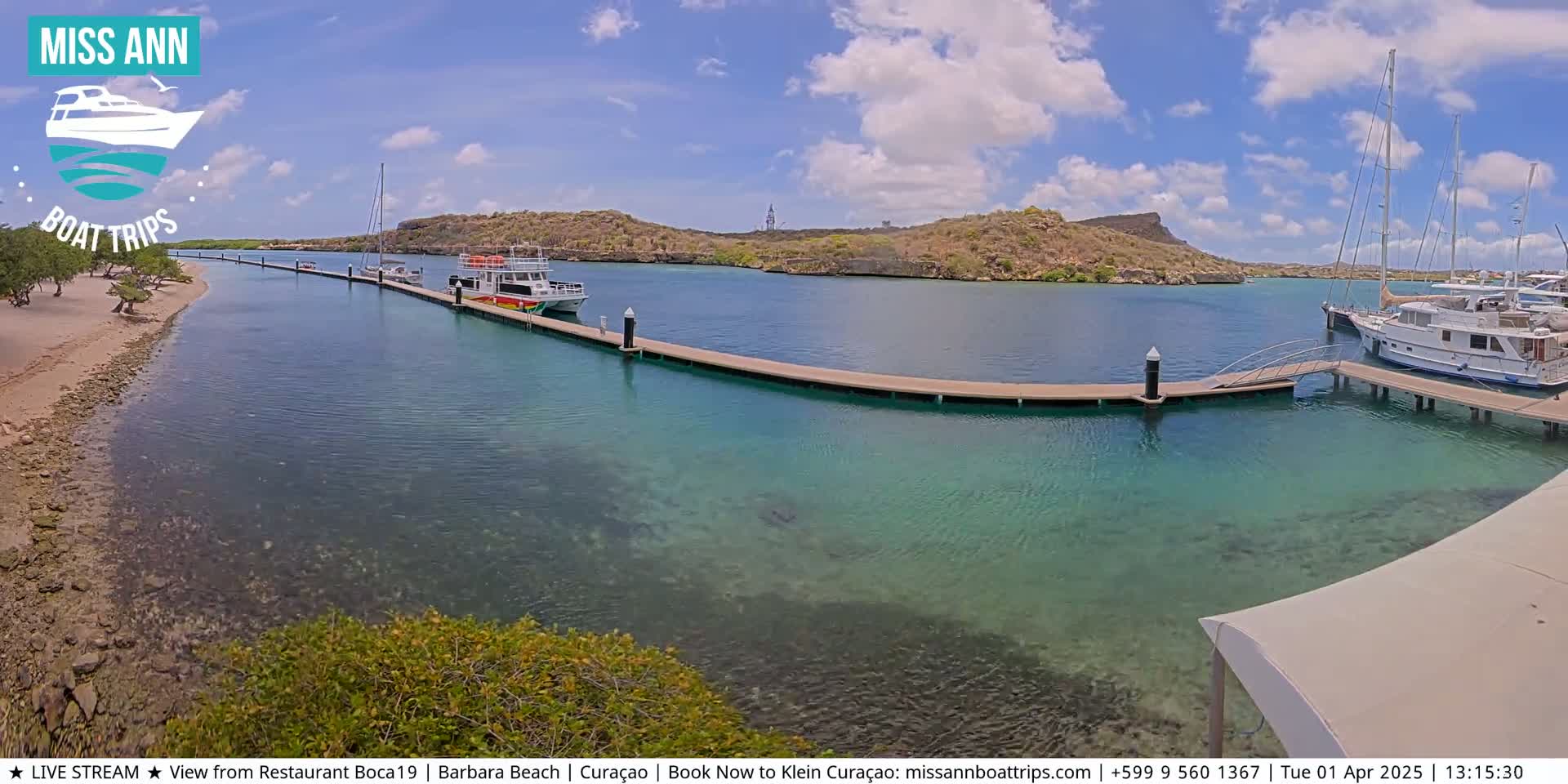 A sunny day shows a calm, clear turquoise body of water with several boats docked at a long wooden pier next to a sandy beach and arid hills.