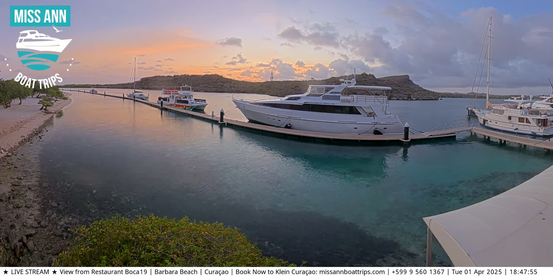 Several boats are docked at a marina during a calm sunset, with clear, shallow turquoise water.