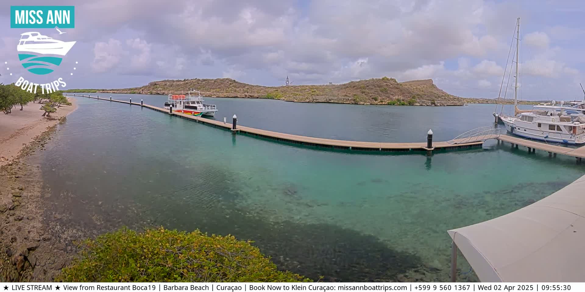 A calm, partly cloudy day shows a marina with several boats docked at piers extending into clear, shallow turquoise water, with a sandy beach and low vegetation visible in the foreground.