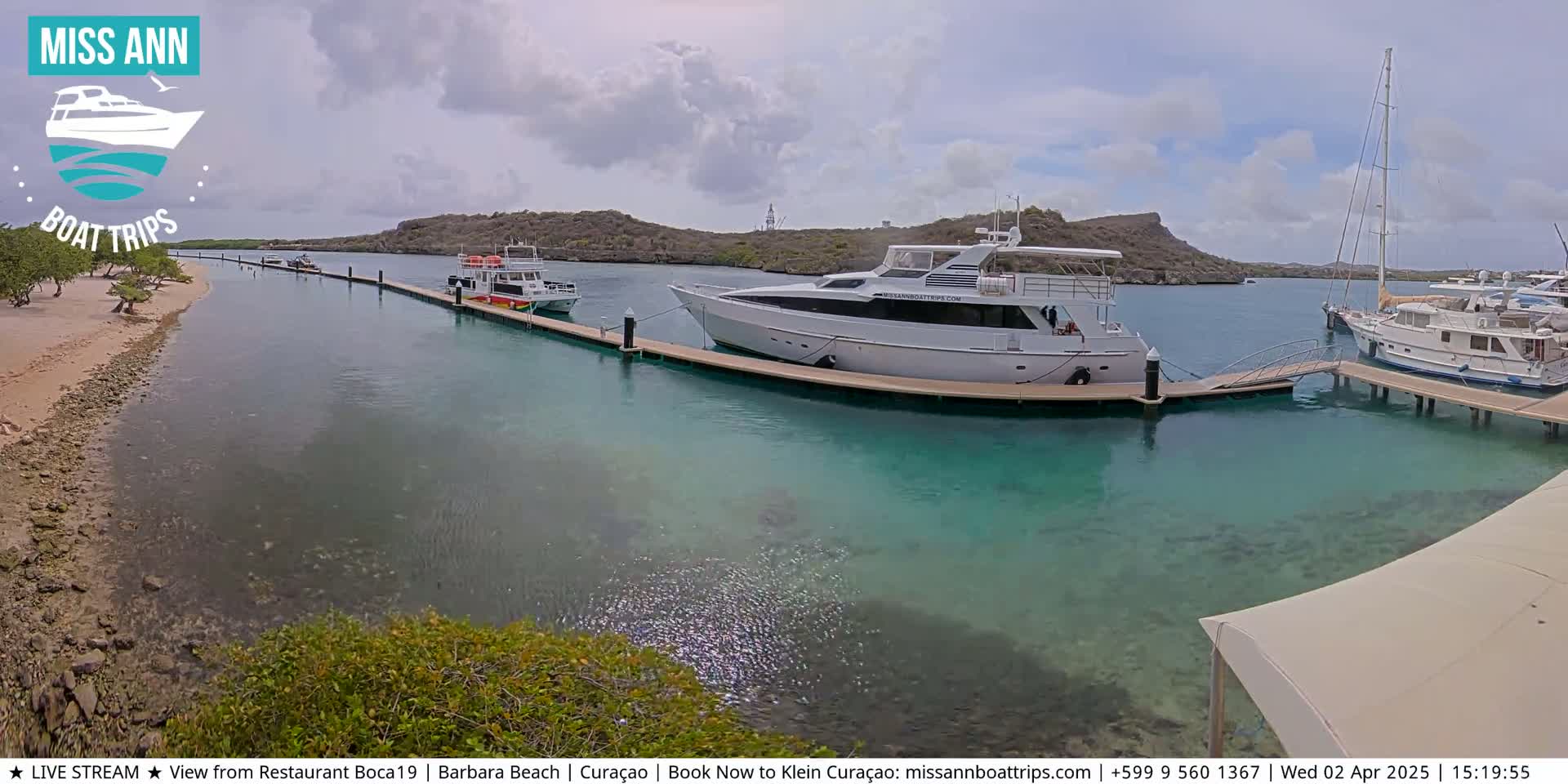 Several boats are docked at a marina on a partly cloudy day, with clear, shallow turquoise water visible around the docks.