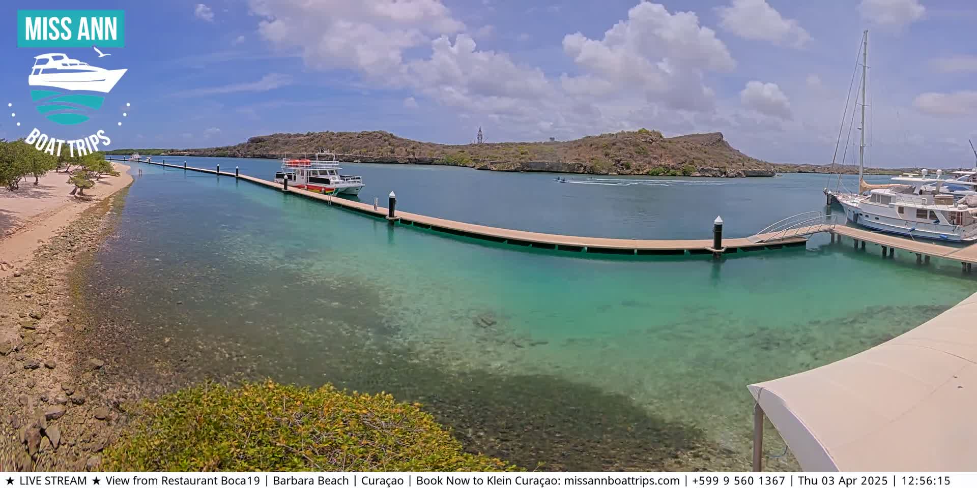 A sunny day shows a marina with several boats docked at piers, clear turquoise water, and a sandy beach with sparse vegetation.