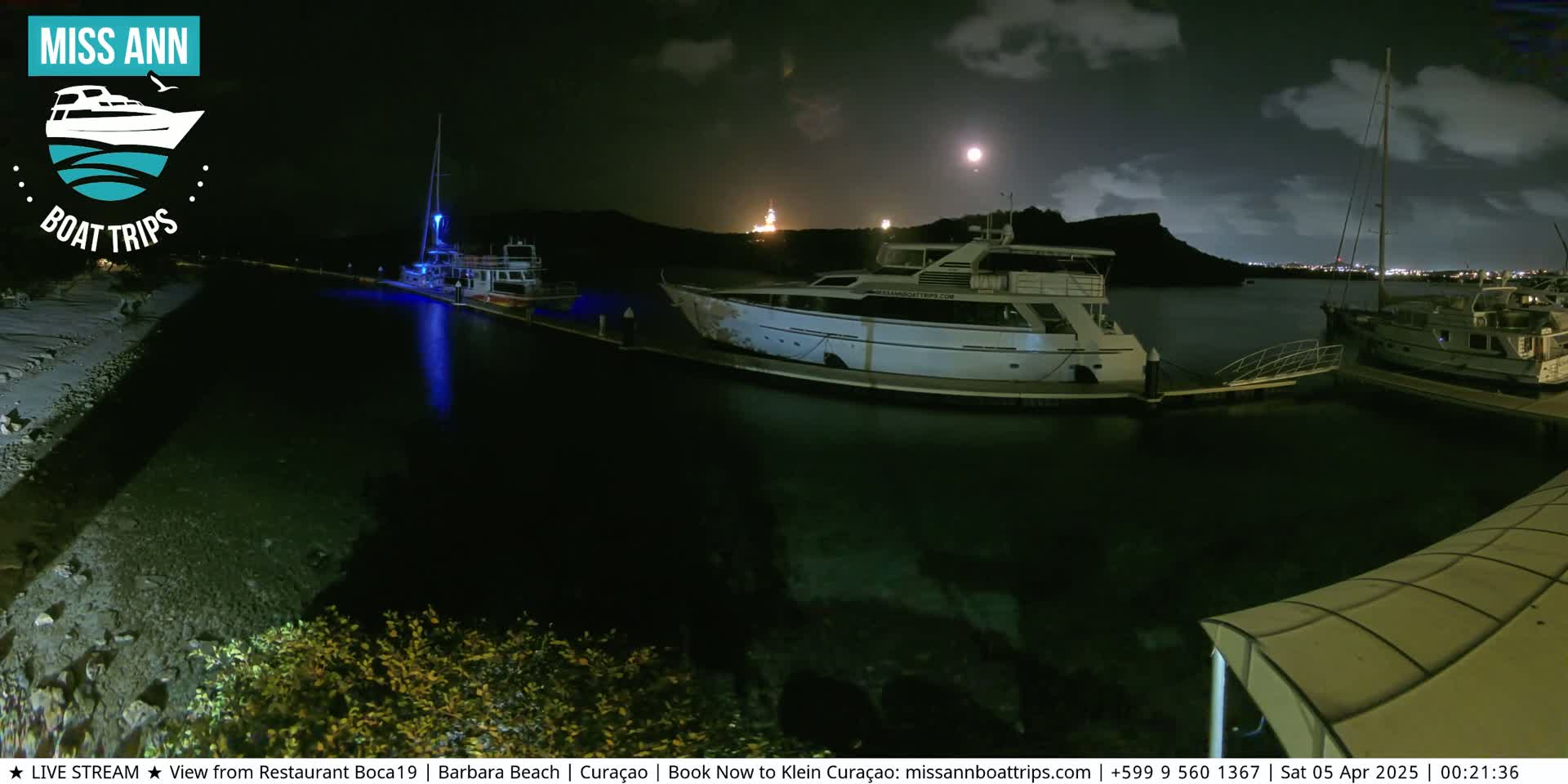 Several boats are docked at a marina at night under a partly cloudy sky, illuminated by a full moon and lights from the boats and shore.